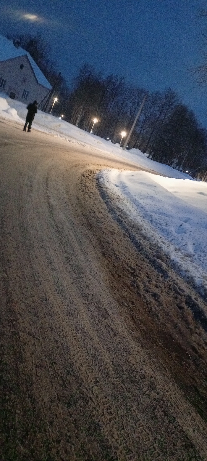The image depicts an outdoor winter scene at dusk or night in Красный Маяк, Russia. A single individual, dressed in dark winter clothing, is seen from behind, walking away from the camera on the left side of a winding road. The road surface consists of compacted slush, dirt, and patches of snow, exhibiting visible tire tracks and faint footprints. To the immediate right of the road, a substantial snow bank is present, with additional snow accumulated along the left edge. A line of streetlights with warm, yellowish-orange illumination extends into the background, casting light onto the road and snow. On the far left, a light-colored building with a peaked roof and multiple windows is partially visible, appearing through sparse trees. A dense cluster of bare trees forms the background on the right side of the road, contrasting with the dark blue, grainy sky. A faint, elongated light source is visible in the upper left portion of the sky. The overall environment is cold and urban or suburban, characterized by winter conditions and artificial lighting.