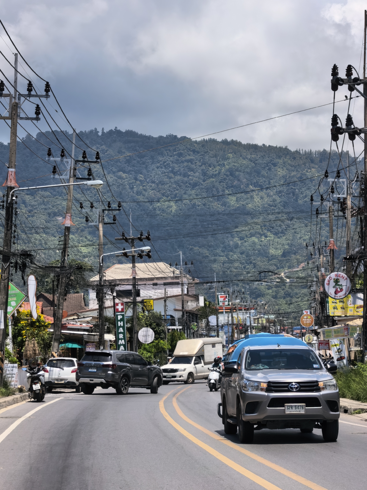 The image depicts a street scene in the city of Ko Samui District, Thailand. A grey pickup truck with a blue tarp covering its cargo is driving on a paved road. Behind it, a white truck and a motorcycle are also visible. On the left side of the road, a white car and a motorcycle are parked. The street is lined with businesses, including a 7-Eleven and a pharmacy. Overhead, a dense network of utility poles and wires stretches across the scene, with a lush, green mountainous landscape in the background under a cloudy sky.