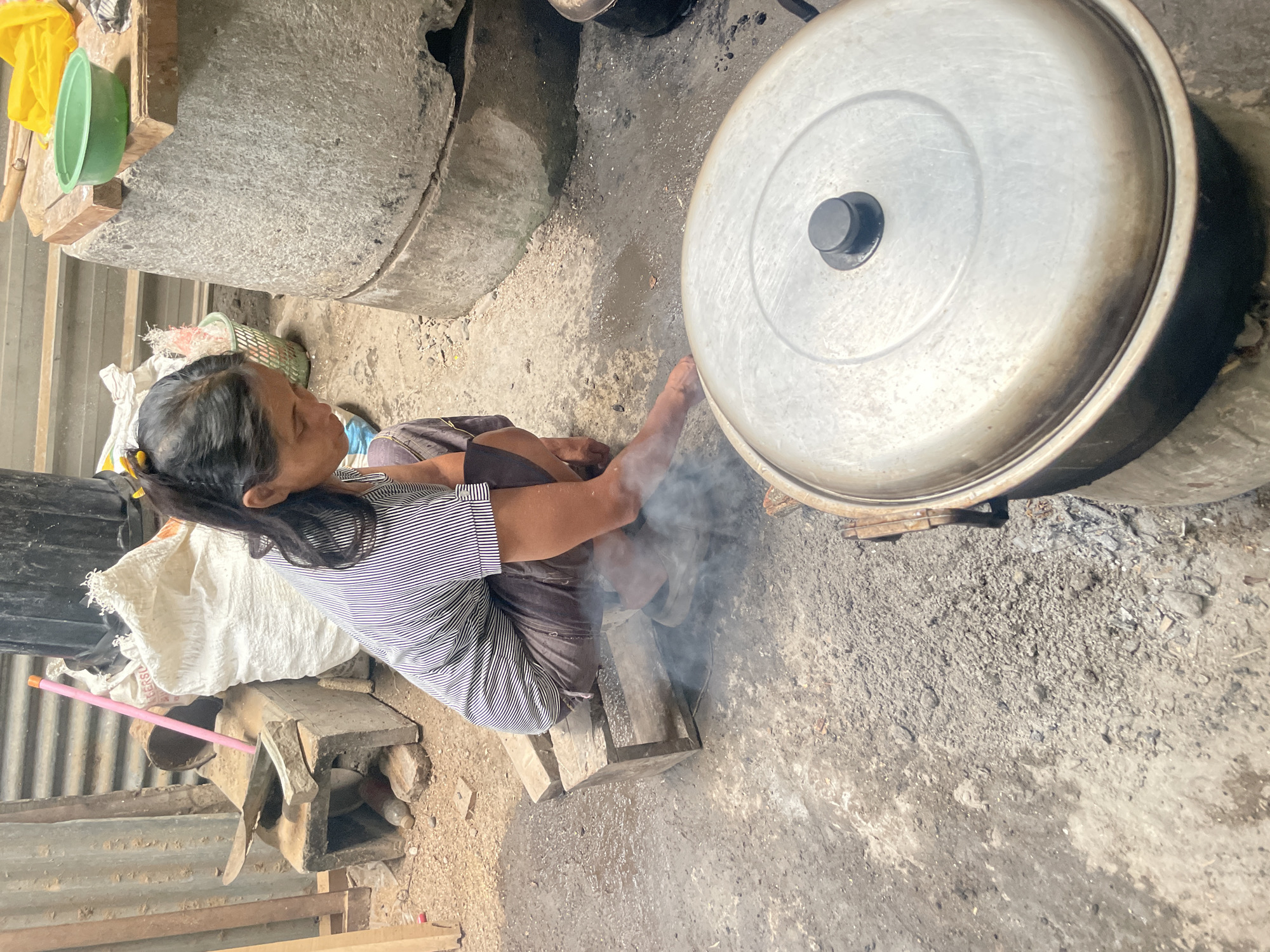 In Parakan, Indonesia, a woman with dark hair tied back in a ponytail, wearing a striped t-shirt and brown trousers, is crouched on a wooden stool next to a rustic outdoor cooking setup. Smoke rises from a large, silver pot with a black lid that is placed on what appears to be a makeshift stove. The ground is a mixture of dirt and gravel, with some scattered debris. In the background, there are various items including a large white sack, a green plastic bowl on a wooden surface, and corrugated metal sheeting. The scene suggests a simple, functional living space where food preparation is taking place.