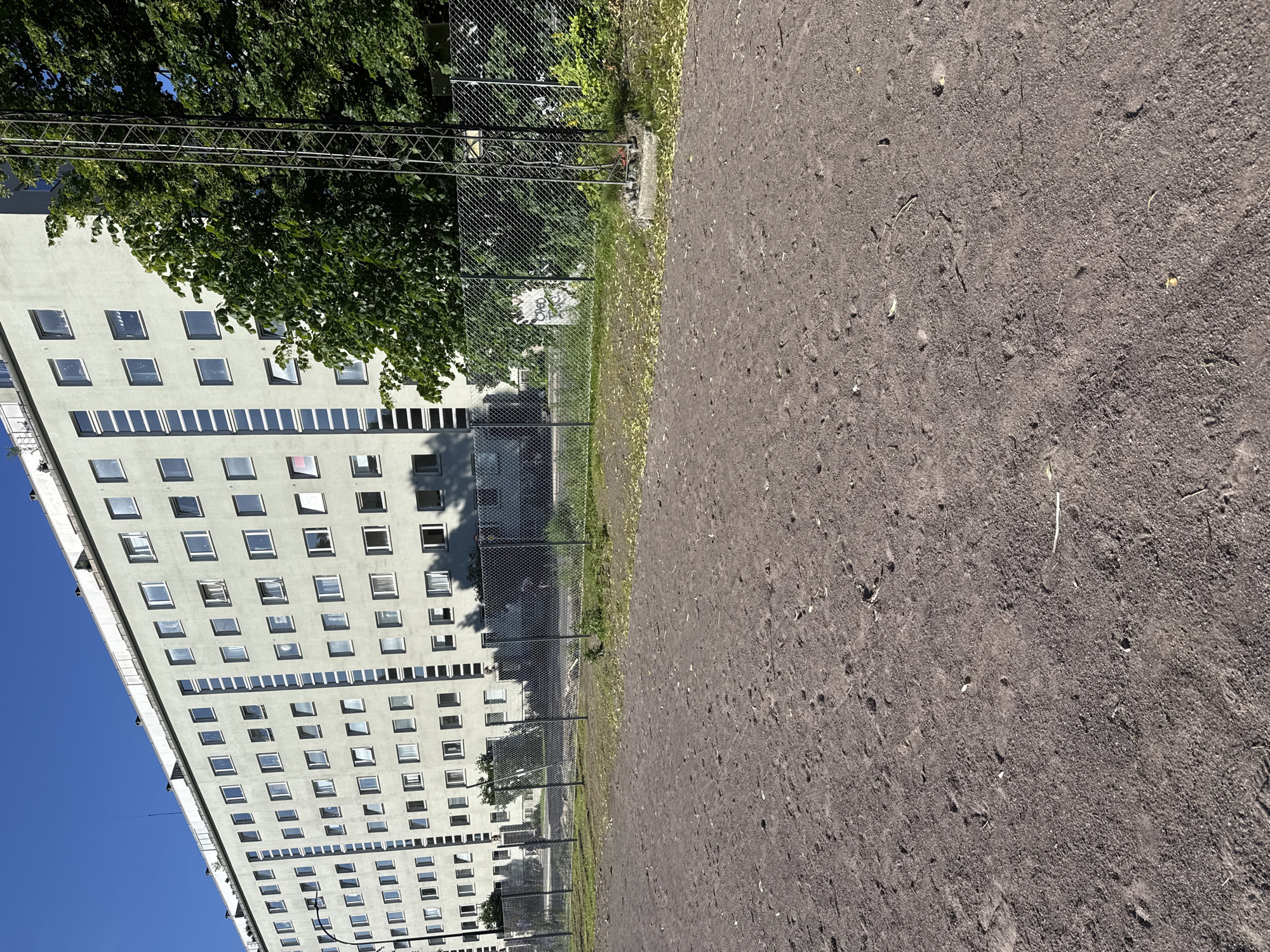 The image depicts a wide, flat, dirt or gravel area in the foreground, with small scattered leaves and debris. A chain-link fence runs horizontally across the middle ground, separating the open area from a grassy strip and more foliage behind it. Beyond the fence and trees, a very long, light-colored apartment building dominates the background. The building has many regularly spaced windows and appears to be several stories high, stretching horizontally across the frame. To the right of the apartment building, a tall metal tower, likely a communication tower, is visible. The sky above is clear and blue. The scene is set during daytime, under bright sunlight, casting shadows. Based on the architectural style of the apartment building and the overall urban setting, this appears to be a residential area within the city of Oslo, Norway. No people are visible in the image.