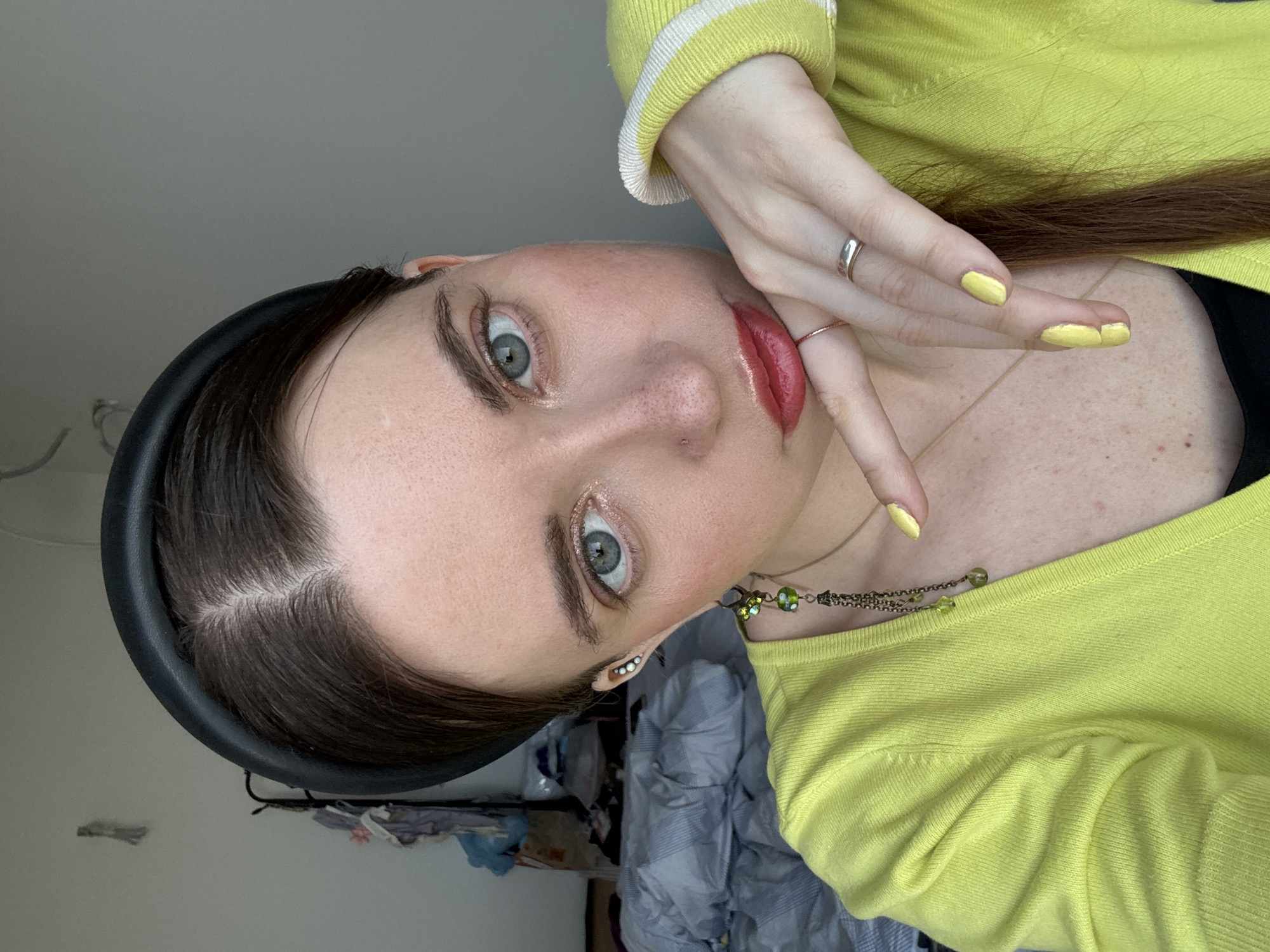 A young woman with brown hair, fair skin, and striking blue eyes is facing the camera. She has a black headband on. She is wearing a light green sweater with white trim. Her fingernails are painted yellow, and she is wearing two rings. She is wearing a necklace with green beads. The background includes a messy room with bedding and some items hanging. The image does not have enough information to determine if the woman or the location are in Moscow, Russia.