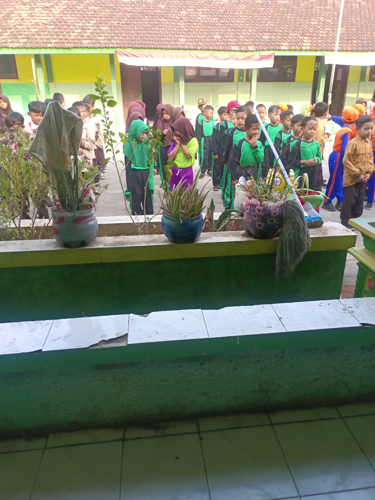 The image depicts an outdoor scene in the city of Sarimulyo, Indonesia, likely at a school, with a group of children, primarily young boys, lined up in rows. The children are dressed in various uniforms, including green and black tracksuits, and some wear hijabs. They appear to be waiting or participating in an activity. In the foreground, there are planter boxes containing plants and decorative items. One planter box holds a plant and a mop. Behind the children, a building with a tiled roof and yellow and green walls is visible. The overall environment suggests a warm climate, with bright sunlight casting shadows.