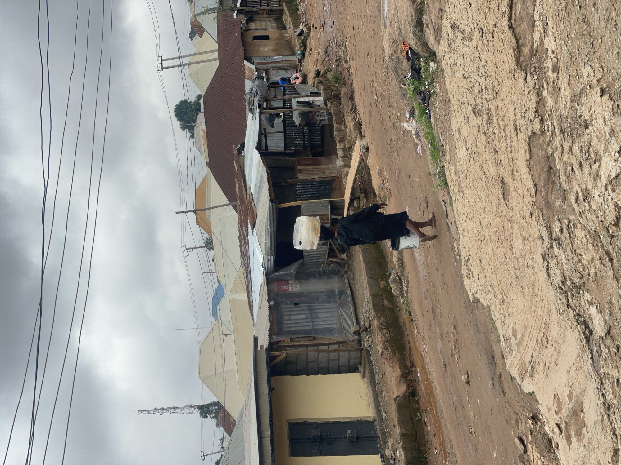 In Kurunduma, Nigeria, a person walks down a dirt road, carrying a large white plastic container on their head and a smaller white bucket in their right hand. The individual is wearing a dark-colored hooded jacket and shorts, with sandals on their feet. The surroundings depict a somewhat cluttered environment with various structures, including buildings with corrugated metal roofs, some appearing to be informal settlements. One building on the left has a yellow facade and a dark metal door. The road is unpaved and muddy in places, with debris and vegetation visible along its edges. In the background, utility poles and wires crisscross the sky, along with a distant telecommunications tower. The sky is overcast with clouds. Several other people are visible in the distance, some near the structures.