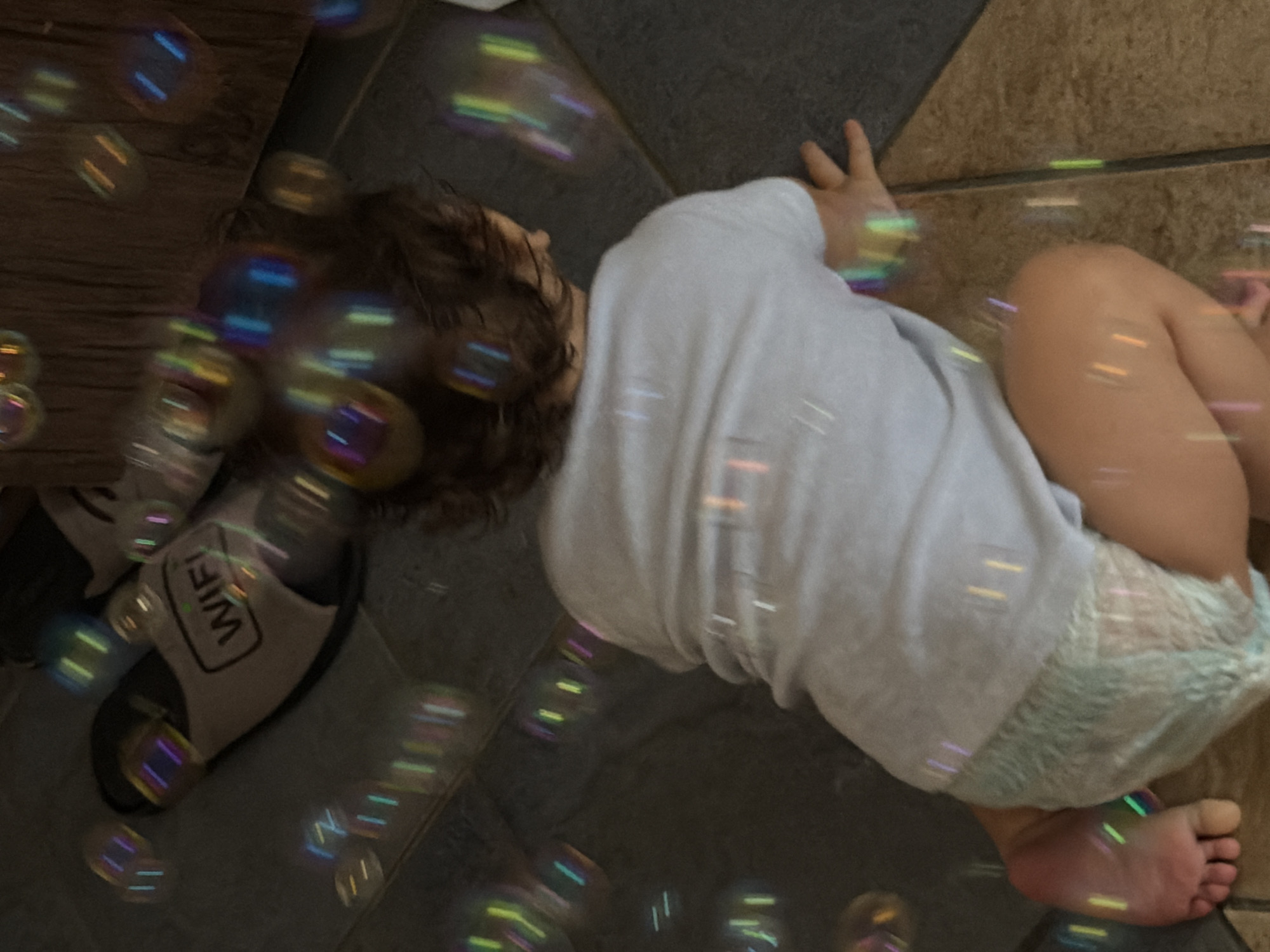 The image shows a baby kneeling on a tiled floor, wearing a diaper and a light-colored t-shirt. Bubbles are floating around the baby, reflecting light. In the background, a pair of slippers with the word "WIFI" printed on them are visible, along with a wooden surface. The scene appears to be indoors, possibly in a home. The image does not contain any elements that would suggest a specific location within Sharm El Sheikh, Egypt.
