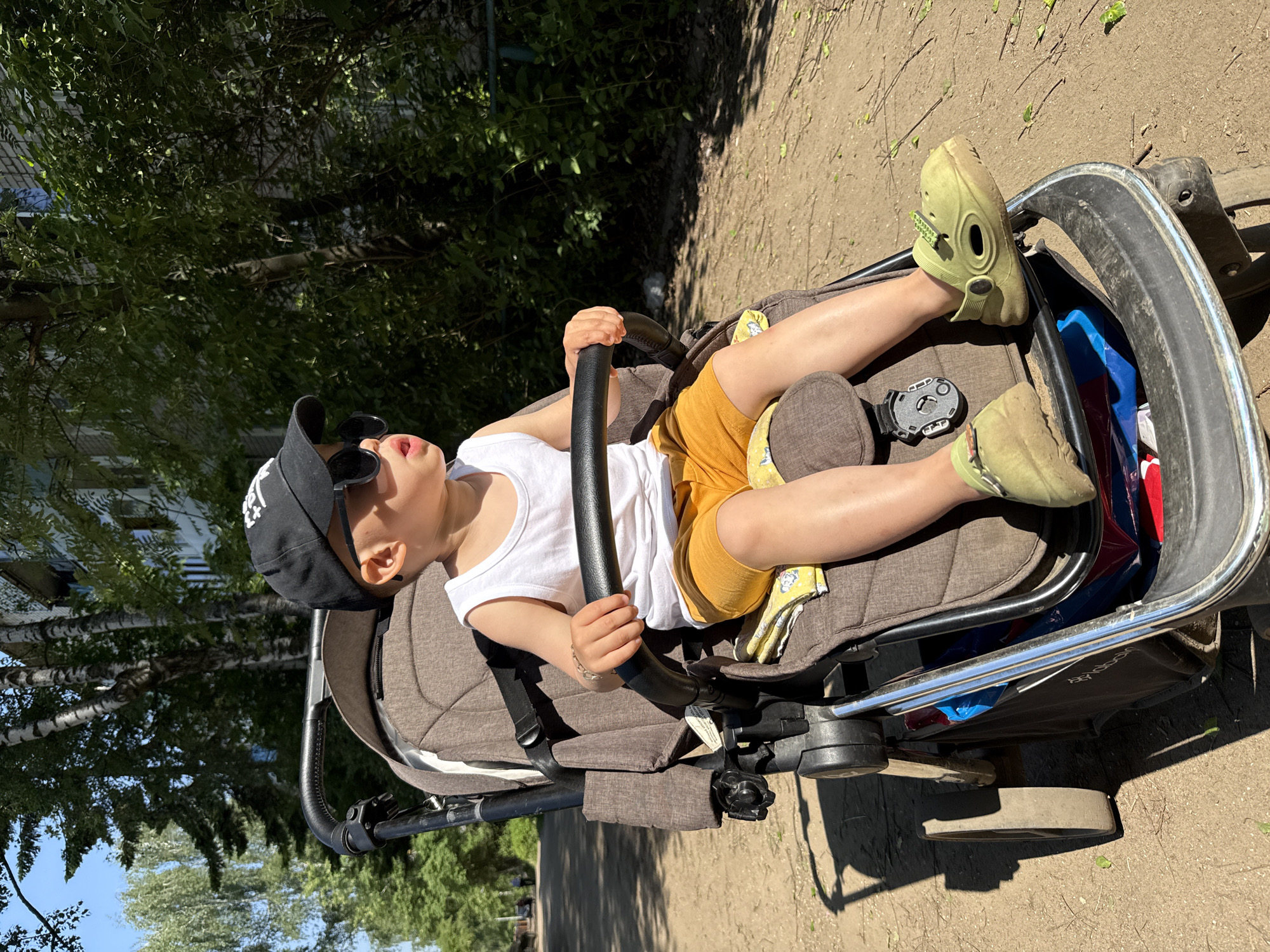 A child wearing a white tank top, yellow shorts, a black baseball cap, and sunglasses is sitting in a stroller. The stroller is on an outdoor surface, possibly a sidewalk or pavement, and is partially in the shade of trees. The child is gripping the stroller's safety bar with both hands. The child is wearing light green shoes. The location is Rusanovo, Russia.