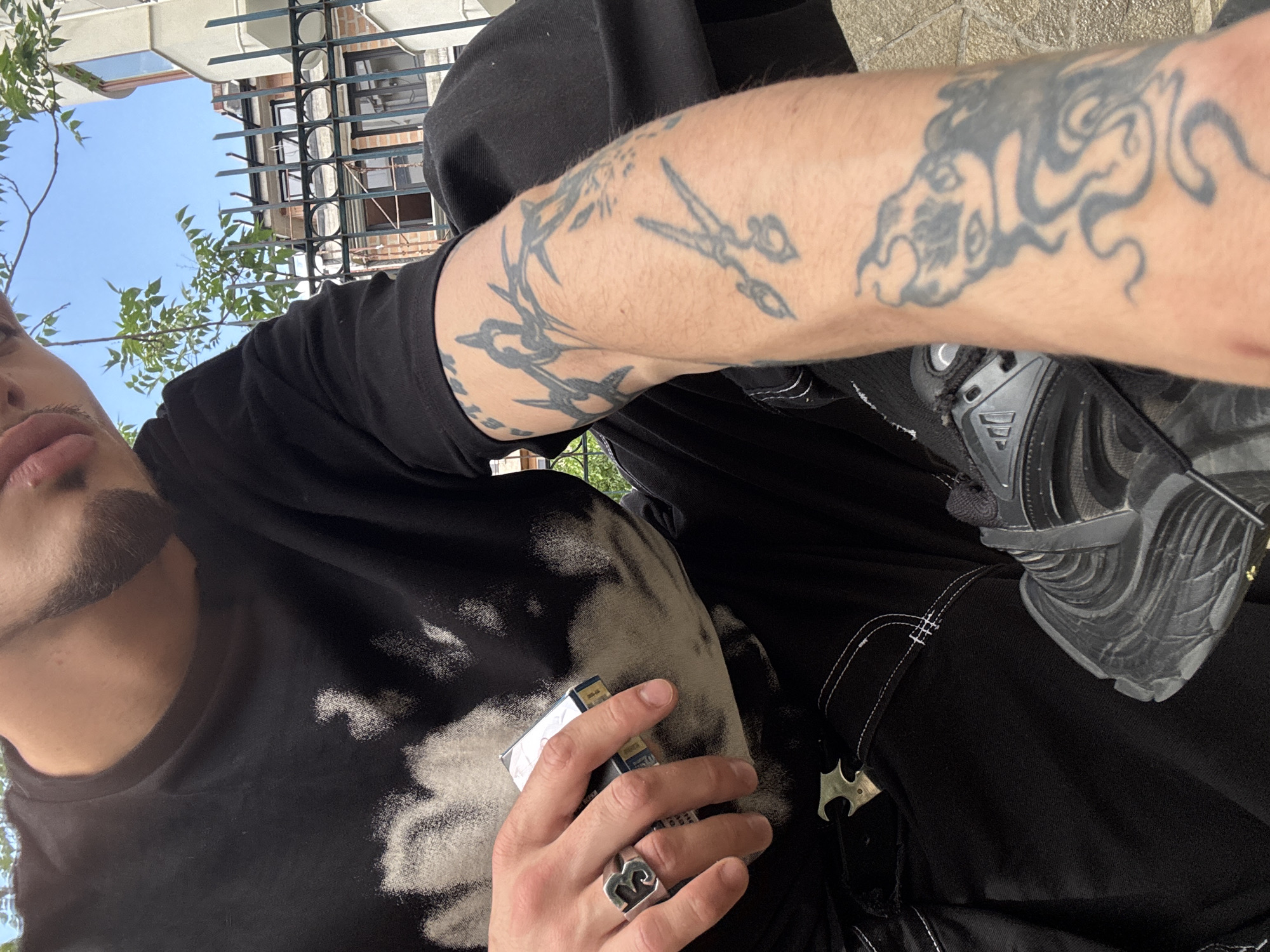 The image is a close-up, low-angle shot of a person in an urban outdoor setting. The person is wearing a black t-shirt with a faded, abstract design on it, and a black hooded garment. Their arm, which is visible on the right side of the frame, is covered in tattoos. They are holding a pack of cigarettes in their right hand, which also features a prominent silver ring with a stylized "B" or similar emblem. Their left hand is visible on their left side, with fingernails that appear unpolished.
The background suggests a bright day, with a clear blue sky visible above some greenery and a building with windows and a metal fence. The person's face is partially visible, showing lips and a goatee. They are wearing black athletic shoes with a complex lacing system and textured soles. The overall mood appears casual and somewhat edgy, with the tattoos and dark clothing contributing to this impression. The time of day is clearly daytime due to the bright sky and the lighting. The presence of a building and foliage suggests an urban or suburban environment. No specific event is depicted; it appears to be a candid shot. No visible text is legible.
