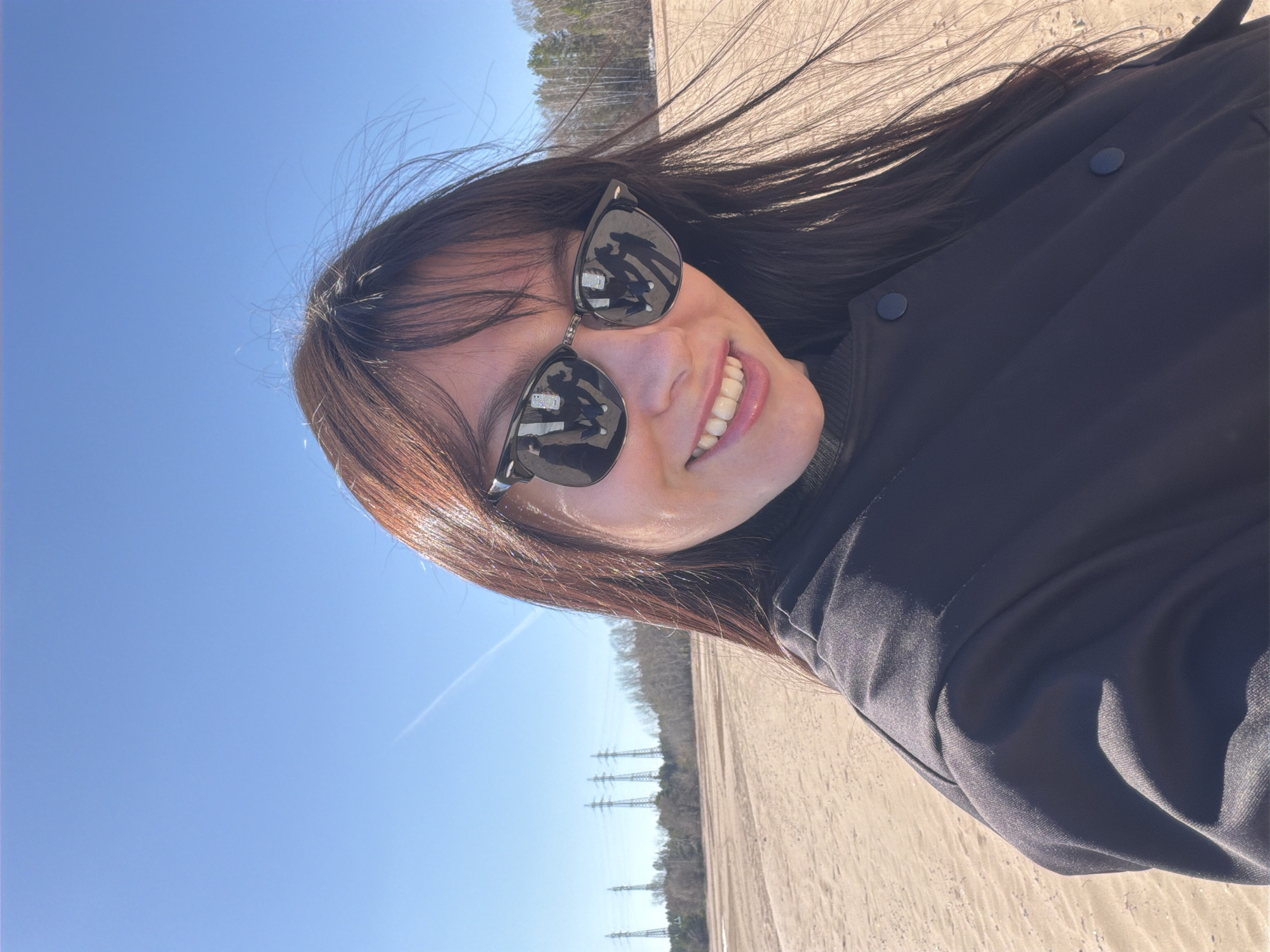 This brightly lit, horizontal selfie captures a young woman smiling broadly outdoors in Chaikovsky, Russia. Her dark, long hair appears to be swept by the wind, framing her face. She wears dark sunglasses that reflect the clear blue sky and parts of the sandy ground. Her wide smile reveals white teeth, conveying a joyful emotion. She is dressed in a dark, possibly black, long-sleeved jacket or coat with two visible dark buttons on the front.
The background features a vast expanse of clear blue sky, with a faint white contrail visible. Below the horizon, a wide area of light-colored, sandy, or dry dirt ground stretches into the distance. A line of bare trees marks the far background, and several tall metal electricity pylons (transmission towers) stand between the trees and the foreground, indicating a somewhat open or rural landscape. The bright sunlight suggests it is a sunny day, likely midday or early afternoon. No visible text is present in the image.