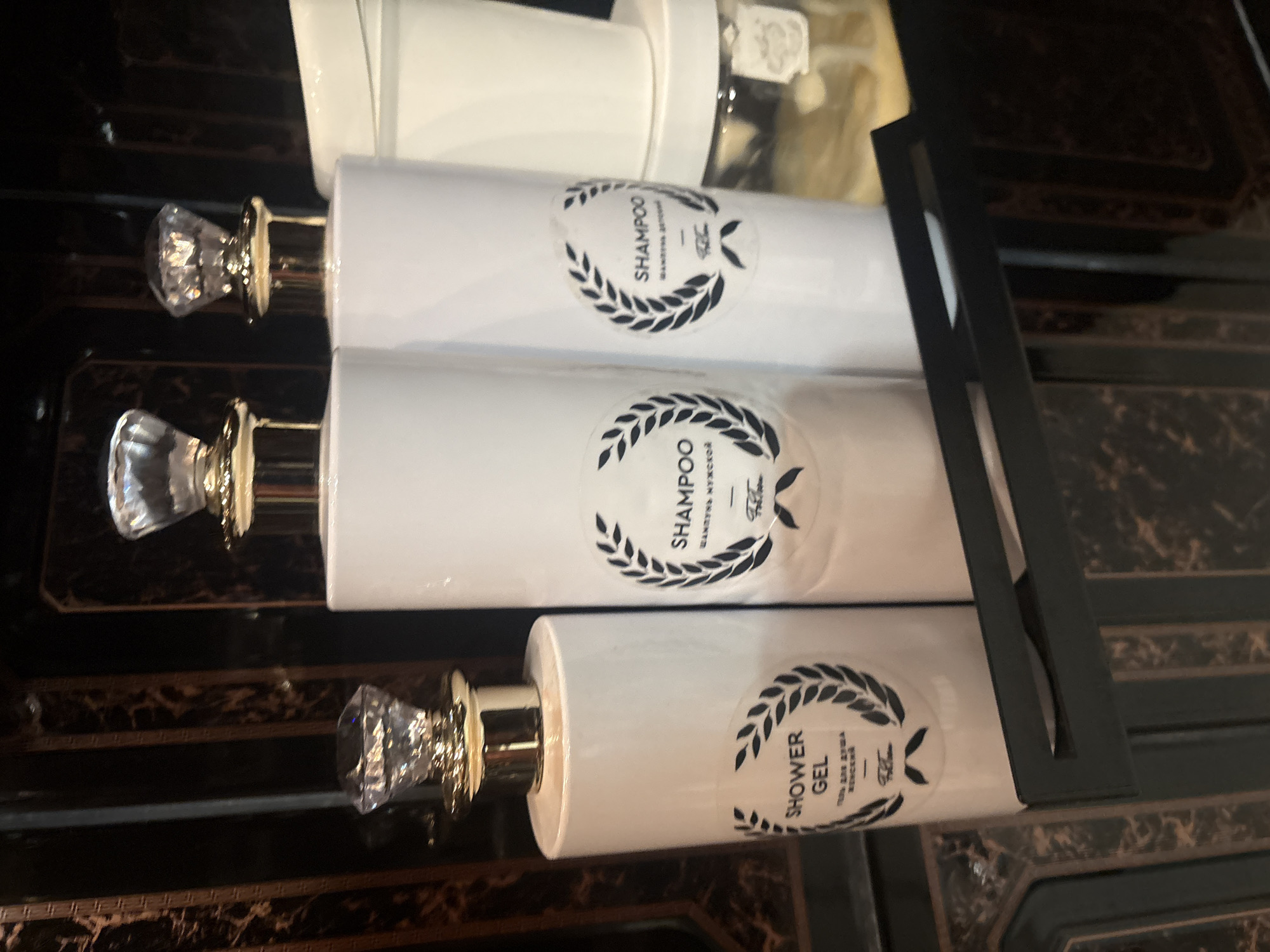 Here's a description of the image:
The image shows three white bottles with a clear liquid inside, arranged horizontally against a dark marble-like surface. Each bottle has a gold-colored pump with a crystal-like knob. The top two bottles are labeled "Shampoo," with text in Russian underneath. The bottom bottle is labeled "Shower Gel" with text in Russian underneath. All three bottles have a wreath-like design surrounding the text. The bottles appear to be part of a set, contained within a black frame or display.
Given the Russian text, the origin is most likely Krasnoyarsk, Russia.