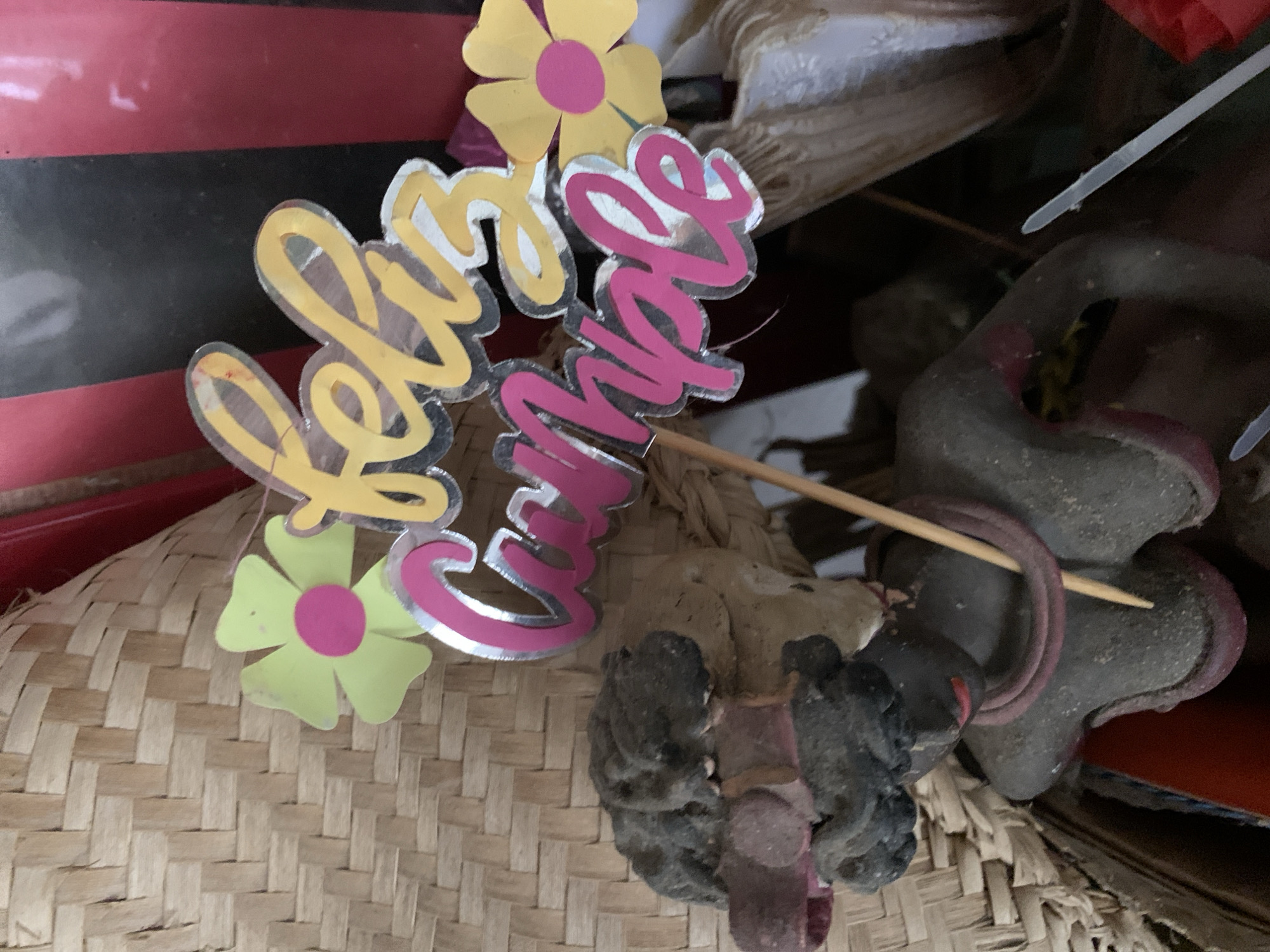 The image shows a close-up of a woven basket with a "Feliz Cumple" (Happy Birthday) cake topper stuck into it. The cake topper is made of yellow and pink lettering with silver edging, and two flowers are attached. Next to the basket is a gray and red ceramic object that appears to be part of a figurine. The background is cluttered with other objects, including some fabric and what appears to be a striped box. The image does not provide information about the location in Santa Ana, Venezuela.
