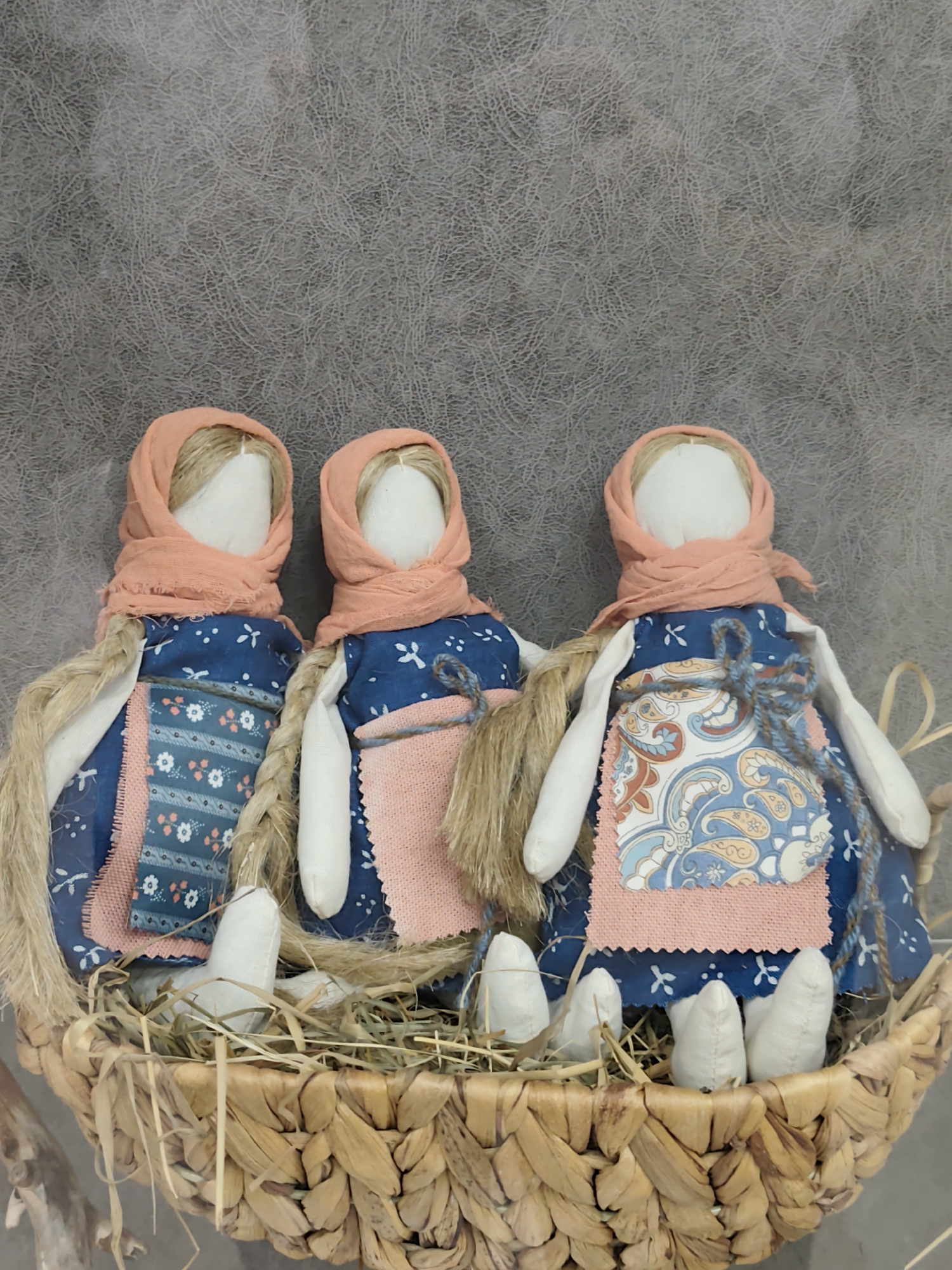 Three traditional Russian folk dolls, known as motanka dolls, are arranged in a woven basket filled with straw. The dolls have white fabric bodies and heads, each adorned with a peach-colored headscarf. They are dressed in blue fabric dresses with white floral patterns and an additional pink apron-like layer. Their arms are simple white fabric tubes, and their legs are pointed white fabric shapes peeking out from the basket. Two of the dolls have blonde yarn braided into pigtails. The background is a textured gray surface. The scene is set indoors, with no discernible outdoor elements or specific Izhevsk landmarks visible. The dolls appear to be decorative items rather than actively engaged subjects.