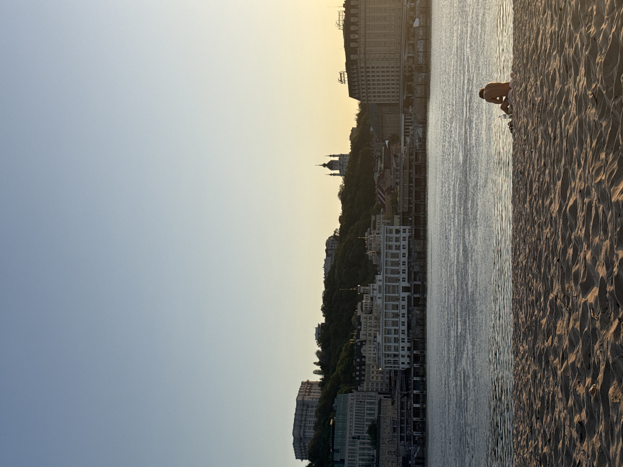 The image depicts a cityscape along the Dnieper River in Kyiv, Ukraine, taken during the early morning or late afternoon. In the foreground is a sandy beach, with a person (adult male) crouched near the water's edge. The river flows between the beach and the city. The city has a mixture of old and modern buildings. The sky is clear, with a gradient from blue to a light yellow hue, indicating the angle of the sun.