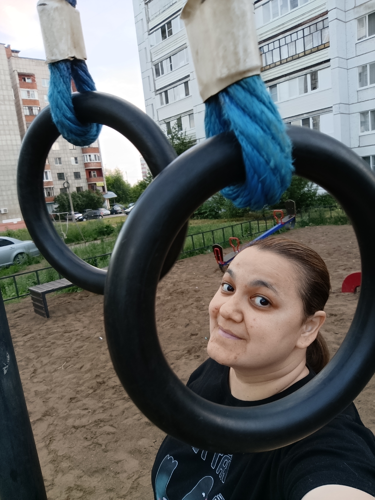 In a Chaikovsky, Russia, playground, a woman with brown hair pulled back poses for a selfie, framed by the black rings of gymnastic equipment. The rings, suspended by thick blue ropes wrapped around white cylindrical fixtures, occupy the foreground. Behind her, a sandy playground surface is visible, dotted with a wooden bench and a seesaw. Apartment buildings, characteristic of Chaikovsky's urban landscape, rise in the background, their facades suggesting a residential area. Cars are parked along the street bordering the playground. The sky is overcast, indicating a temperate climate.