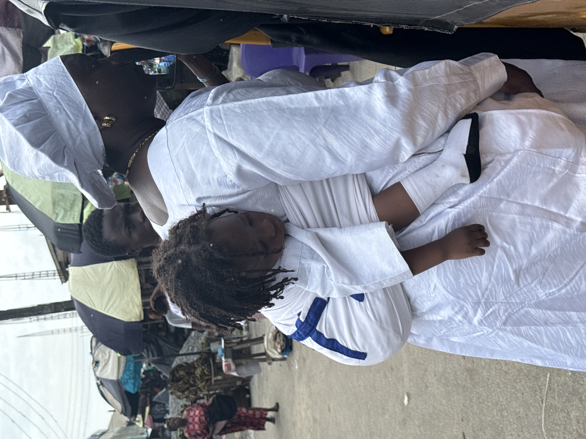 A woman, wearing a white headwrap, gold earrings, and a matching white outfit, carries a child on her back in a white sling with blue trim. The child is asleep, with its head resting on the woman's shoulder. The woman's face is turned away from the camera, and she appears to be walking through a market. In the background, there are market stalls with umbrellas, a pile of bananas, and other people. The ground is paved with asphalt. The scene is set in the city of Ado, Nigeria, likely a bustling market environment.