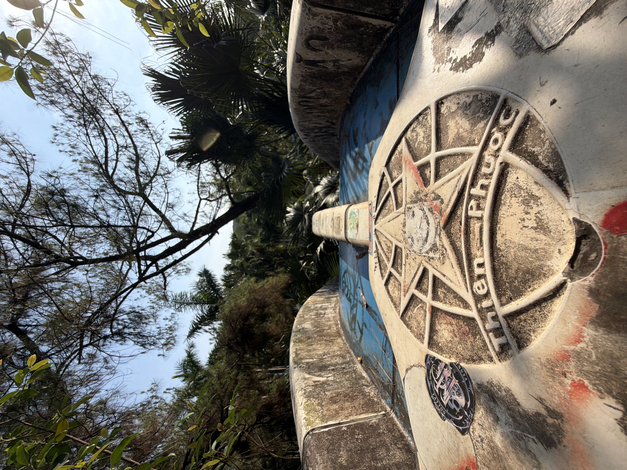 The image shows a close-up of a weathered, likely concrete, structure. The structure features a compass-like design with the words "Thien Phuoc" inscribed in a circle. The design is worn, with some staining and apparent graffiti. The structure is set against a backdrop of lush greenery, including various trees and foliage. The scene is located in Thủy Xuân Ward, Vietnam, as indicated by the inscription on the structure. The view is from a low angle, looking upwards. The sky is partially visible through the trees, appearing bright. There are stickers present on the structure.