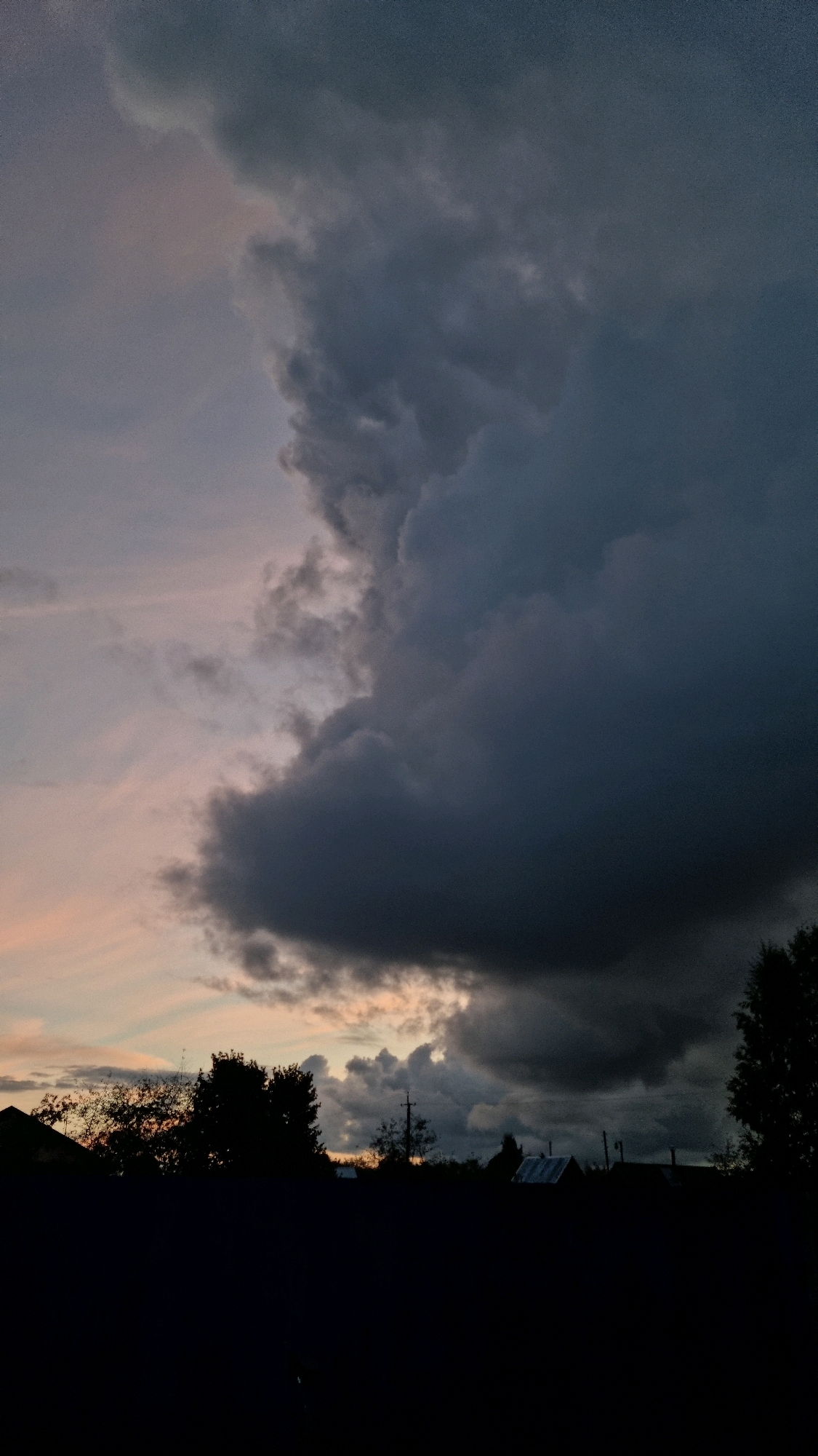 The image captures a dramatic sky over the city of Vysokoe, Russia, just before sunset. A large, dark storm cloud dominates the right side of the frame, casting a heavy shadow. To the left, lighter clouds are tinged with the soft oranges and pinks of the setting sun, creating a stark contrast with the storm. Silhouetted against the sky are the dark shapes of trees and rooftops, indicating a rural or semi-urban setting. A single utility pole is visible in the distance, and the foreground is entirely dark, obscuring any ground-level details. No people or specific activities are discernible.