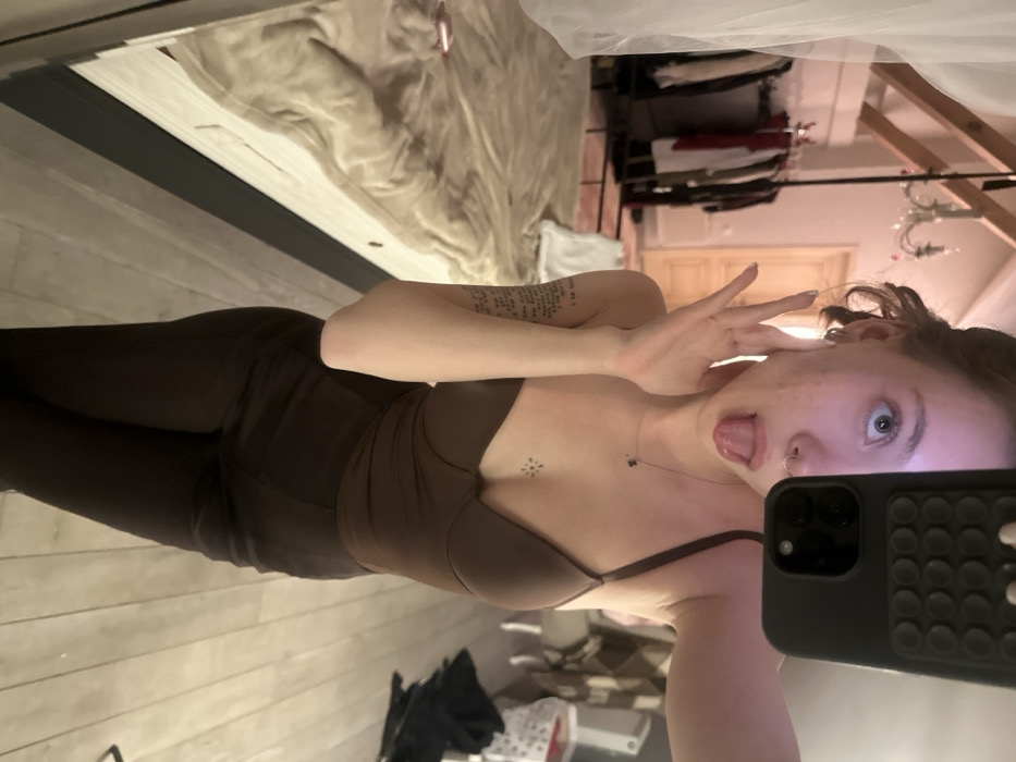 A mirror selfie captures a woman in brown athleisure wear. Leggings and a matching top accentuate her figure. She playfully sticks out her tongue and makes a hand gesture near her face, while holding a phone. Tattoos are visible on her arm and chest. The background reveals a bedroom setting with a bed, clothing rack, and chandelier. The angle and reflection create a distorted perspective.
