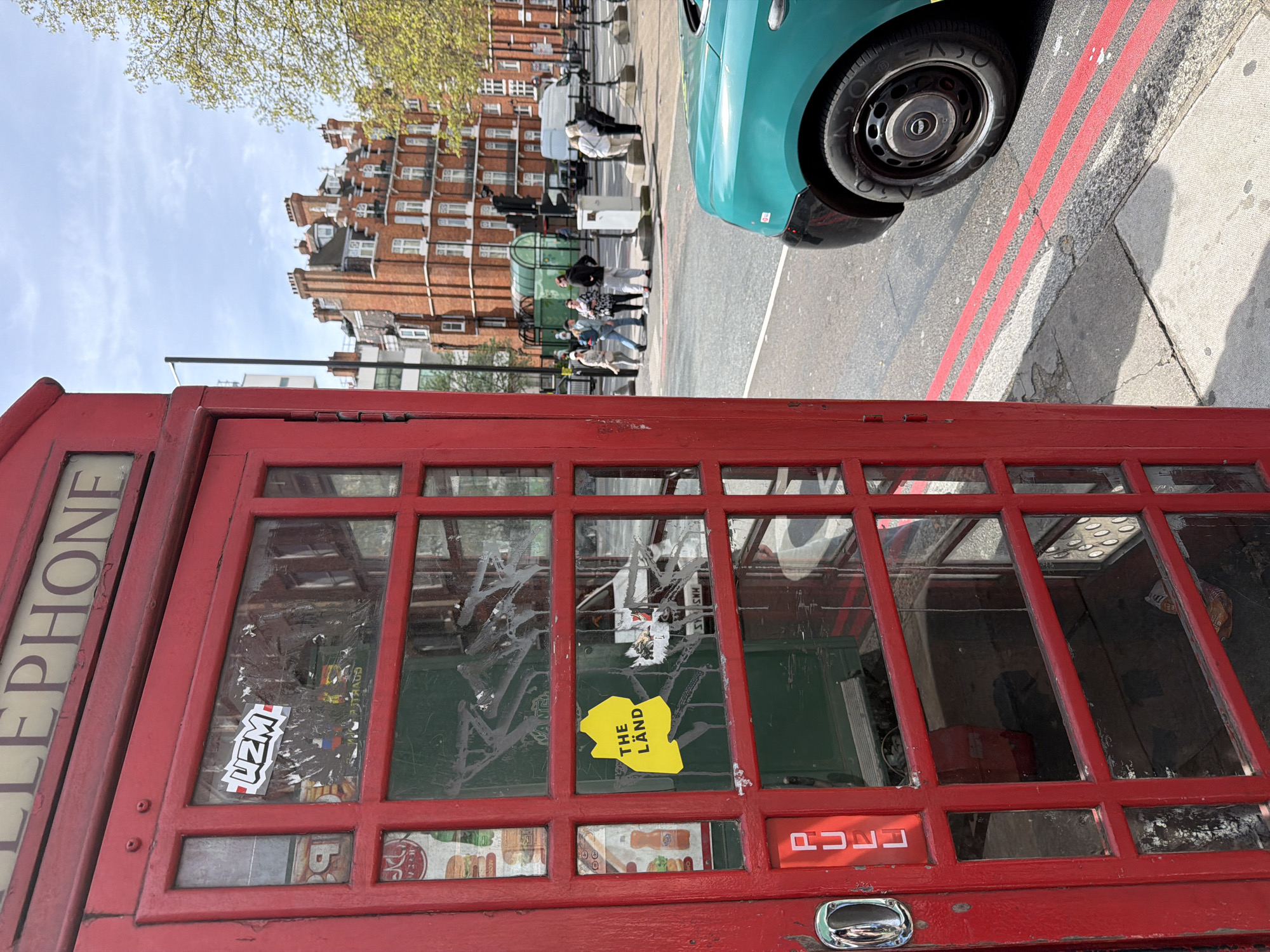 A red telephone booth dominates the foreground. The word "TELEPHONE" is vertically printed on the left side of the booth. The glass windows of the booth are covered with stickers and graffiti. A yellow sticker with the words "THE LAND" is visible on one of the panes. Through the glass, the interior of the booth appears cluttered. Behind the booth, a London street scene unfolds. A line of people are waiting, likely at a bus stop or queue. A turquoise car is parked along the curb. The architectural style of the buildings in the background is traditional London brick. The sky is a clear blue with a few scattered clouds. The overall impression is of a typical London street scene featuring a classic telephone booth.