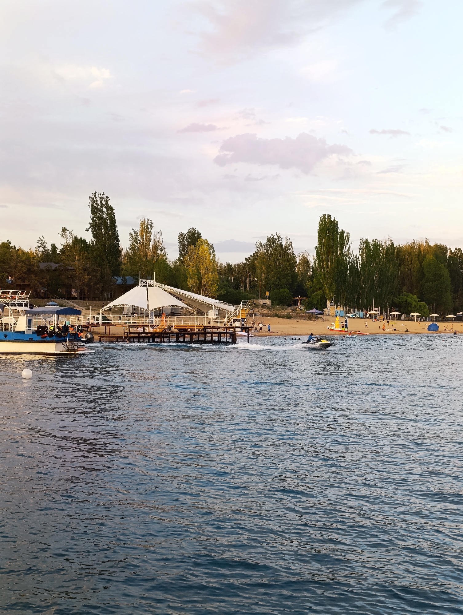 This image depicts a waterfront scene, likely a beach resort area, in the city of Bayet, Kyrgyzstan. In the foreground, the blue water of a lake or sea is visible with moderate ripples. On the right side of the frame, a jet ski is being ridden by a person, creating a spray of water. Further back, on the sandy shore, numerous people are present, some walking, others relaxing, with beach umbrellas and tents scattered about. A long pier with a white canopy structure extends into the water, and a boat is docked on the left side. A line of trees forms the backdrop, with some buildings visible behind them. The sky is overcast with soft clouds, and the overall lighting suggests it is late afternoon or early evening.