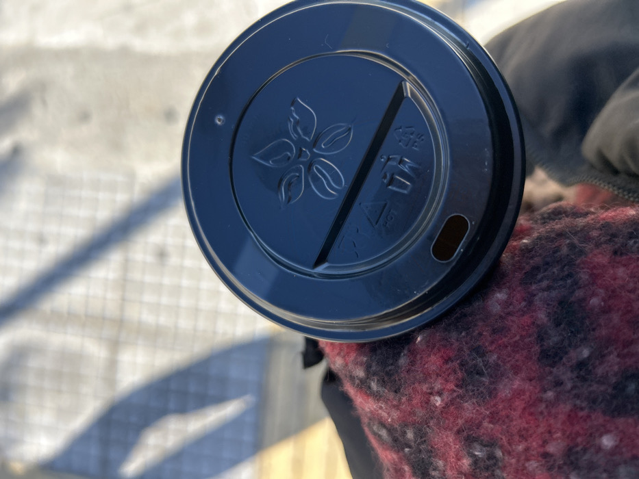 The image presents a close-up, high-angle view of a black plastic coffee cup lid, held by an unseen individual. The lid prominently features a central embossed design depicting a stylized coffee plant or floral pattern, complete with multiple leaves and what appear to be coffee beans. To the right of this design, a clear triangular warning symbol is visible, accompanied by the word "HOT" and a small icon of a steaming cup, indicating the contents are heated. An oval-shaped drinking opening is present on the right edge of the lid.
The cup lid rests against a textured fabric, primarily dark red and black with a checkered pattern, likely part of a jacket, sweater, or scarf worn by the person holding the cup. A darker, solid-colored garment, possibly a sleeve, is partially visible beneath the checkered fabric.
The background, softly blurred, reveals a light-colored paved surface with a subtle grid pattern, strongly illuminated by bright sunlight. Distinct shadows are cast across the ground, suggesting a clear, sunny day, likely during the morning or early afternoon in Nikea, Greece. The scene implies the activity of someone carrying or enjoying a hot beverage outdoors.