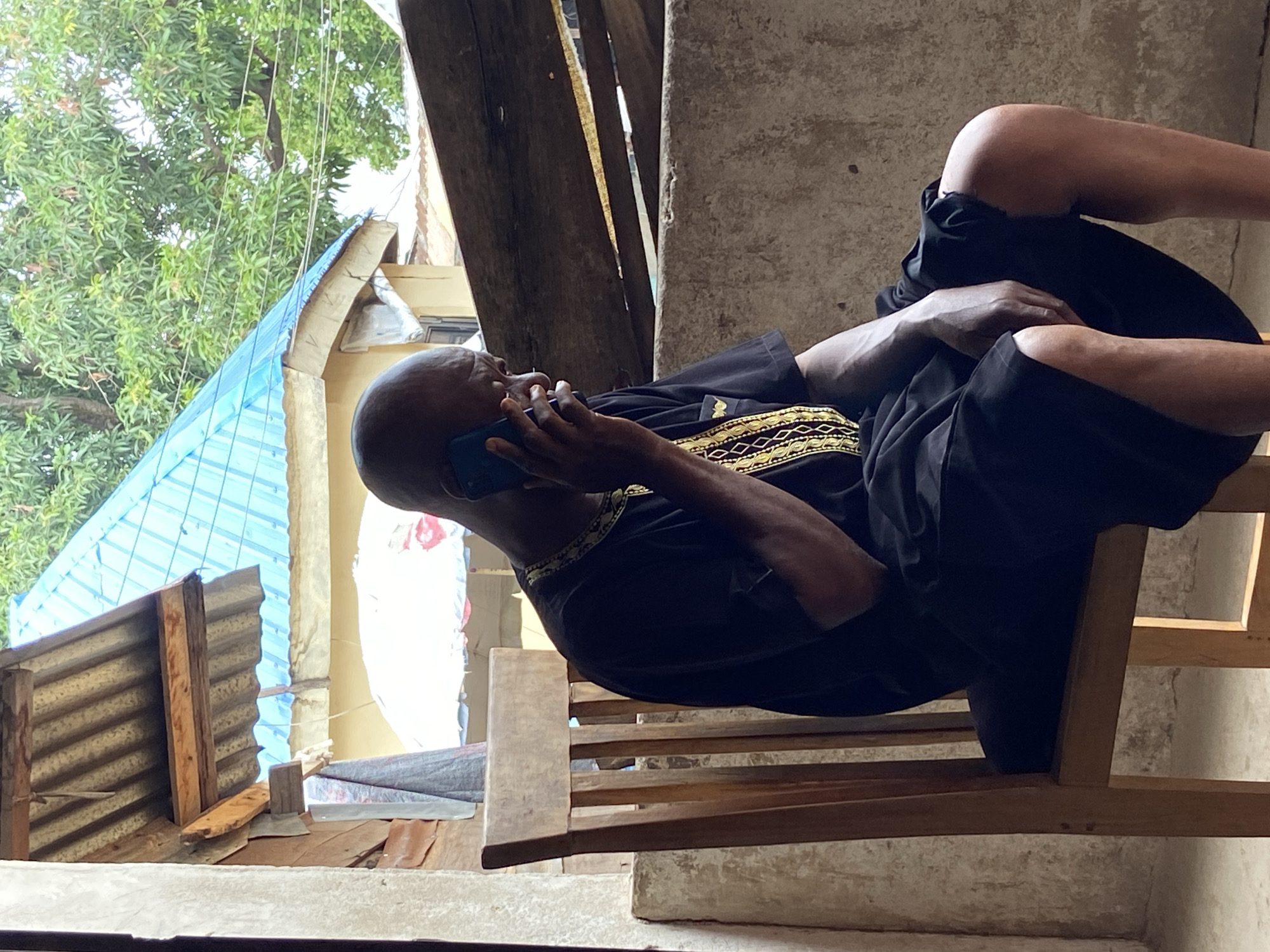 An adult African man is lying on a wooden bench, holding a cell phone to his ear. He is wearing a dark-colored garment with gold embroidery around the neck. The bench is located in an outdoor space, possibly a porch or balcony, in Mararaba, Nigeria. The environment includes corrugated metal structures, a blue-roofed building, and trees, typical of the urban landscape. The man appears to be engaged in a phone conversation.