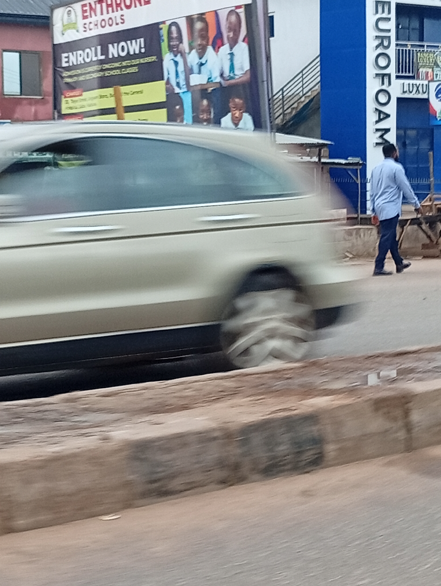 The image depicts a street scene in Sabon Tasha, Nigeria. A light-colored car is moving quickly, creating a blurred effect. A man in a light blue shirt and dark pants is walking on the sidewalk. In the background are advertisements for "Enthrone Schools" and "Eurofoam". The environment appears to be an urban area with buildings and signage.