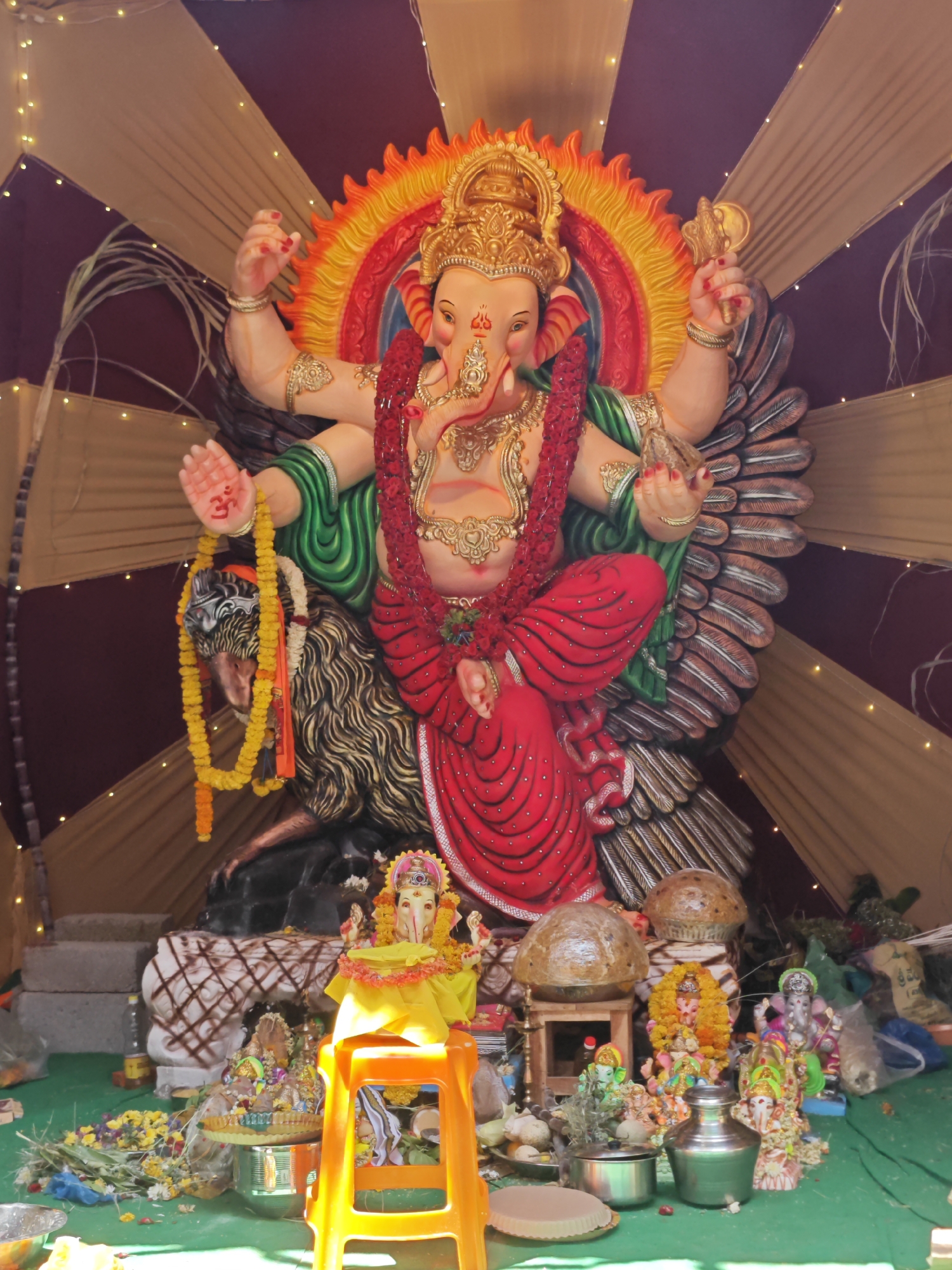 This image captures a vibrant display of Ganesh idols, likely during a religious festival in Puttaparthi, India. The central and largest idol depicts Lord Ganesha seated on a lion, adorned in a red garment, green shawl, and gold jewelry. His hands hold various symbolic objects. A smaller, yellow-clad Ganesh idol is positioned on an orange stool in the foreground, flanked by other smaller Ganesh idols of various colors and sizes. The idols are surrounded by offerings, including garlands of yellow flowers, greenery, and what appear to be stacked coconuts or rounded objects. The backdrop consists of draped fabric in alternating bands of maroon and beige, illuminated by numerous small, warm-toned fairy lights. The ground is covered with a green cloth. The scene suggests a devotional setting, possibly a temporary altar or shrine set up for worship.