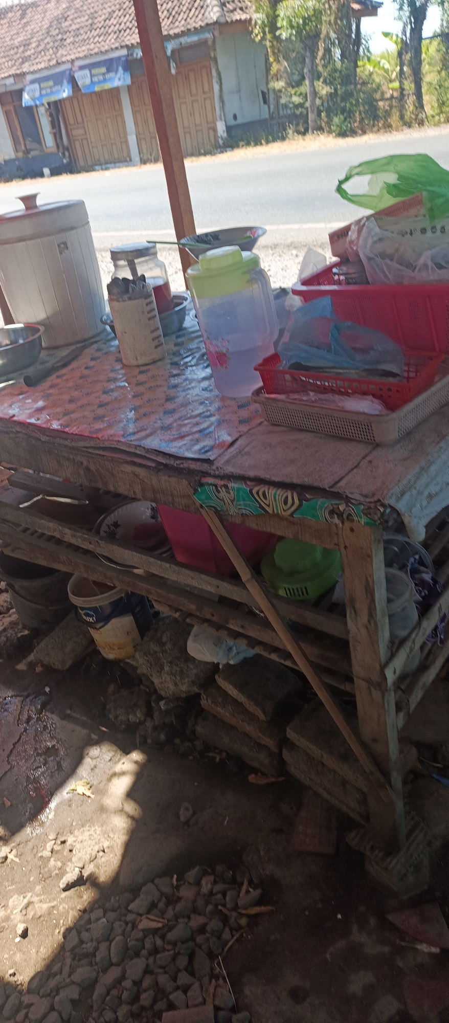 An outdoor scene at what appears to be a roadside food stall in Jatisari, Indonesia. A wooden table, covered with a patterned cloth, displays various items including a large plastic water jug with a green lid, a metal canister filled with utensils, and a red plastic basket containing items wrapped in plastic. To the left, a large white plastic container and a metal bowl are visible. The table is set up outdoors, with a road and buildings in the background. Below the table, a collection of objects, including buckets, rocks, and debris, are scattered on the ground. The ground is uneven and appears to be made of dirt and gravel, with some puddles of water. Sunlight casts shadows across the scene.