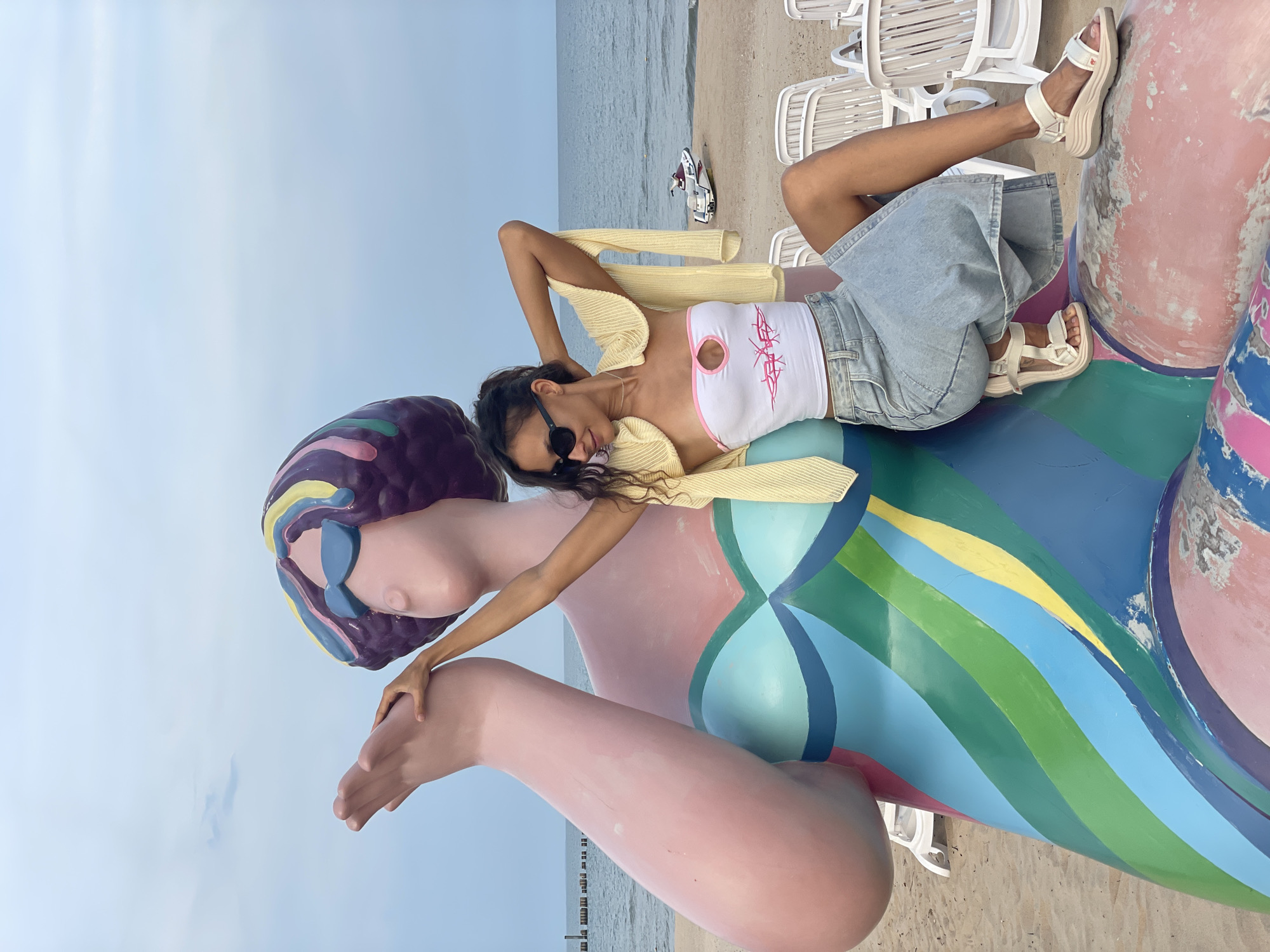 A young woman with long dark hair and sunglasses sits on a large, colorful sculpture of a woman on a beach. The sculpture is pink with a light blue bodice and a multicolored stripe pattern on its lower body and arms. The woman is wearing a white tube top with pink trim and a cut-out, a light yellow cardigan draped over her shoulders, a light blue denim skirt, and white sandals. She is posing with her arms behind her head and one hand resting on the sculpture's arm. The beach is sandy, with lounge chairs and the ocean in the background. The sky is overcast.