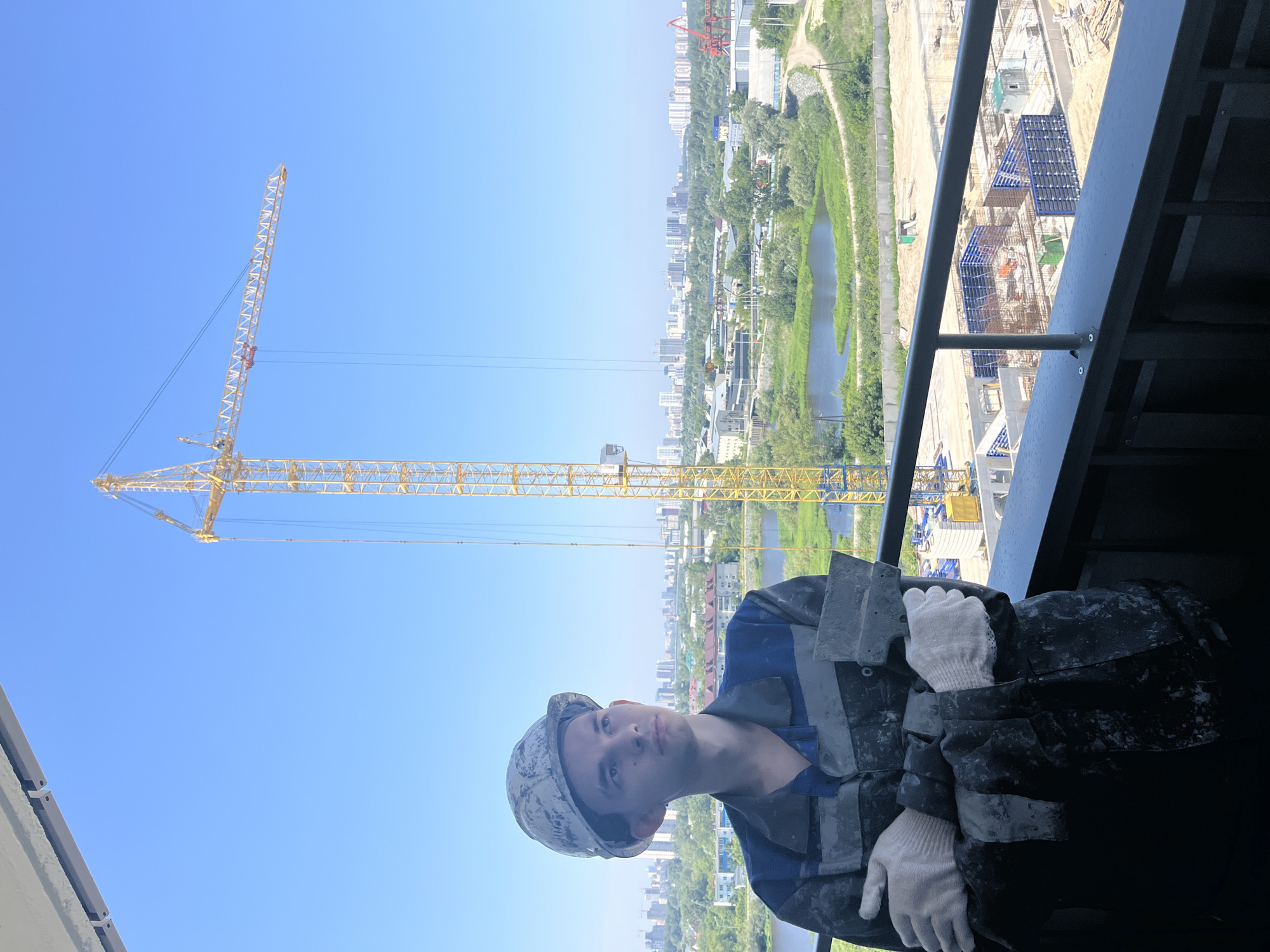 In Tyumen, Russia, a young man stands on a balcony, looking towards a cityscape dominated by a large yellow construction crane. He wears a dirt-stained work uniform and a white hard hat, with white gloves on his hands, one of which holds a trowel. The background reveals a construction site with building foundations, a river, and a distant skyline of multi-story buildings. The sky is clear blue.