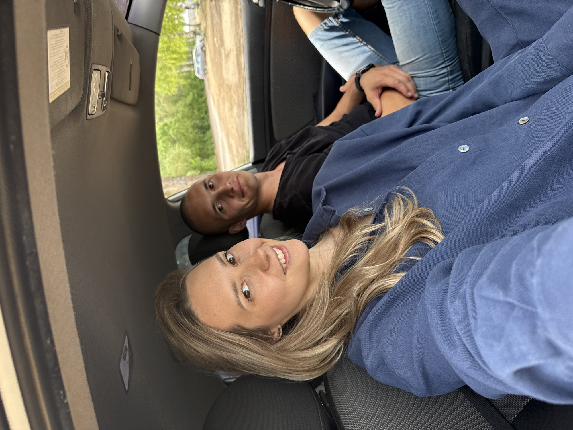 Inside a vehicle, a man and a woman are lying down, apparently in the back seat. The woman is closer to the camera, smiling, and wearing a blue shirt. The man is behind her, wearing a black shirt and looking at the camera. Another person wearing blue jeans is visible in the background. The car is stationary, and a glimpse of the outside shows greenery and a building. There is no information to determine the location is Chaikovsky, Russia from this image.