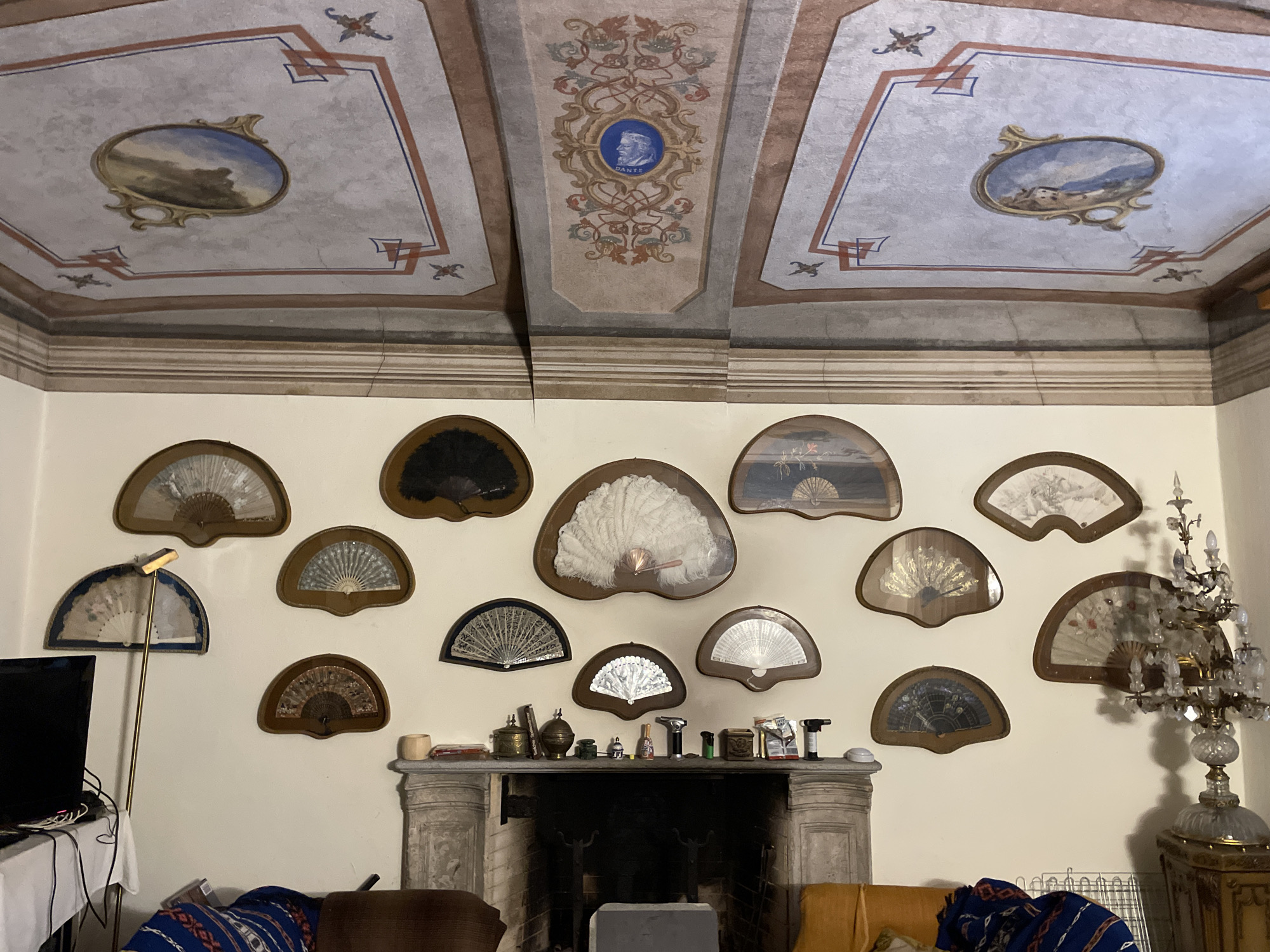 This is a medium shot, eye-level interior photograph of a room, likely in Borgosesia, Italy, featuring a collection of decorative fans displayed on a beige wall above a stone fireplace. The ceiling is ornately painted with a central medallion containing a portrait of Dante. The wall hosts an assortment of fans, varying in size, material, and design, mounted in wooden frames. Some fans appear to be made of lace or fabric with intricate patterns. The fireplace is made of rough-hewn stone and has a dark opening. On the mantelpiece, an array of small objects, including lighters, tins, and decorative items, are arranged. To the left of the fireplace, a black television screen is visible next to a floor lamp with a golden stand. To the right, a crystal chandelier and a gilded cabinet are present. The room also contains furniture, including a dark brown armchair and an orange couch. The overall impression is one of a well-decorated, possibly historical, interior space.