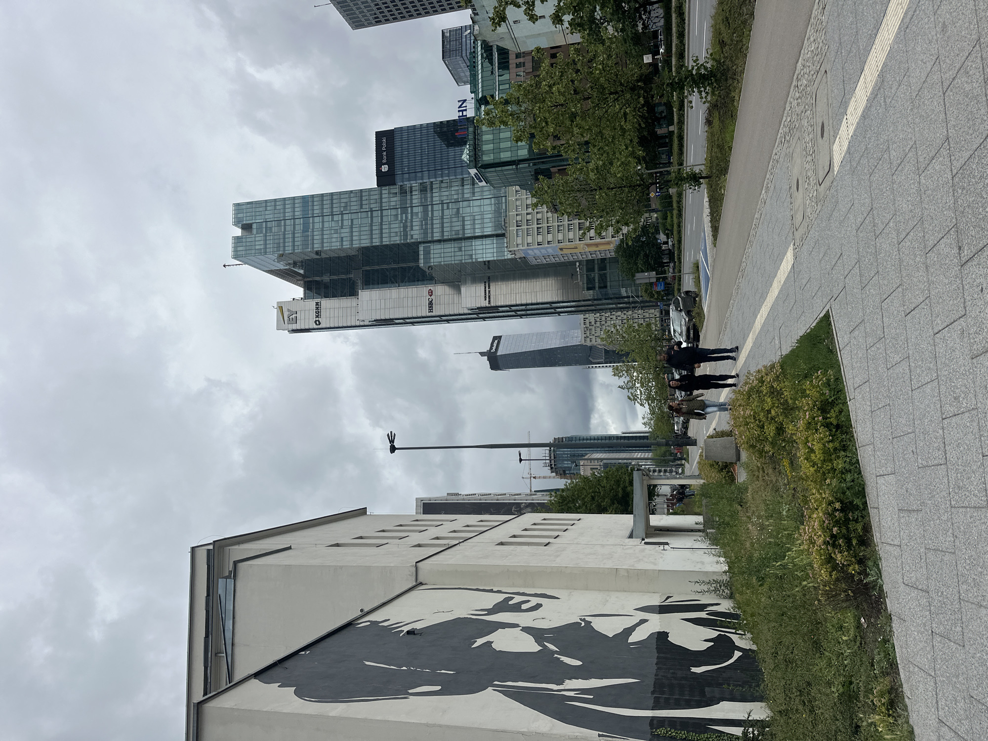 The image depicts a street scene in Warsaw, Poland, featuring a large mural of a person's face on the side of a white building on the left. The mural is rendered in black and white, in a graphic style. Adjacent to the building, a patch of green vegetation and bushes is visible. A paved sidewalk with tactile paving runs alongside the vegetation.
In the background, a modern cityscape unfolds with numerous glass-fronted skyscrapers. Notable buildings include one with the "EY" logo and "Konimpex" text, another with "HSBC" and "Volkswagen Financial Services" signage, and a prominent skyscraper bearing the "Bank Polski" logo. A Deloitte building is also visible further in the distance. A street with cars and a few pedestrians is present. Two people are walking on the sidewalk in the middle ground, and a lamppost with multiple lights stands on the right side of the street. The sky is overcast with clouds. The overall impression is of a contemporary urban environment with a mix of art and modern architecture.