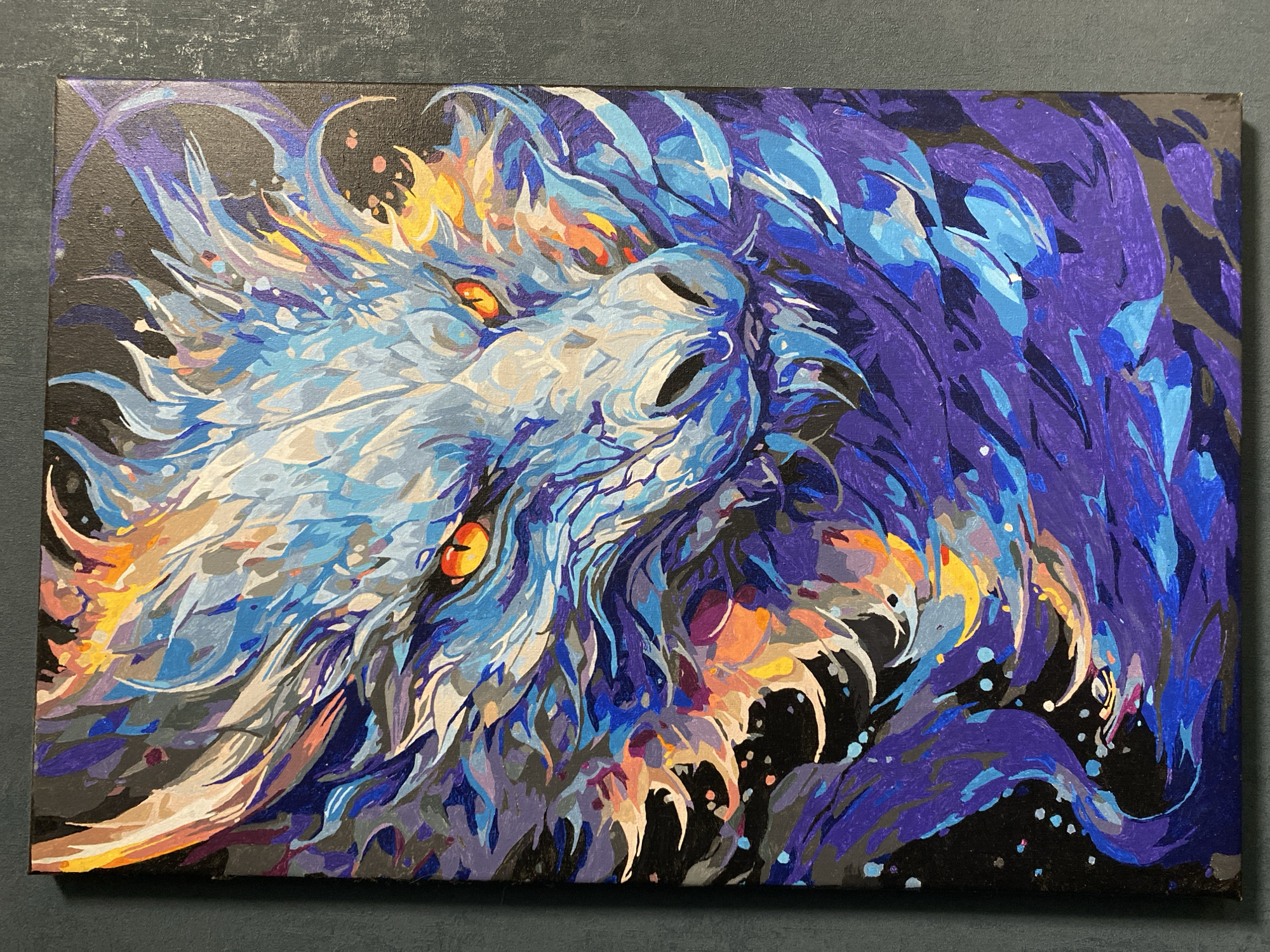 The image is an acrylic painting on canvas depicting a dragon in a stylized, fragmented manner. The dragon's head is prominently displayed, with the left side of its face primarily in shades of white, blue, and gray, accented with orange near the eyes. The eyes are a vibrant orange. To the right, the dragon's mane or scales transition into darker blues and purples, contrasting against a black background speckled with small dots. The composition emphasizes angular shapes and defined brushstrokes, suggesting a paint-by-numbers or similar style. The canvas has a black border and sits against a matte gray surface. The image does not appear to be located in Chaikovsky, Russia, nor does it contain any information that would indicate such a location.