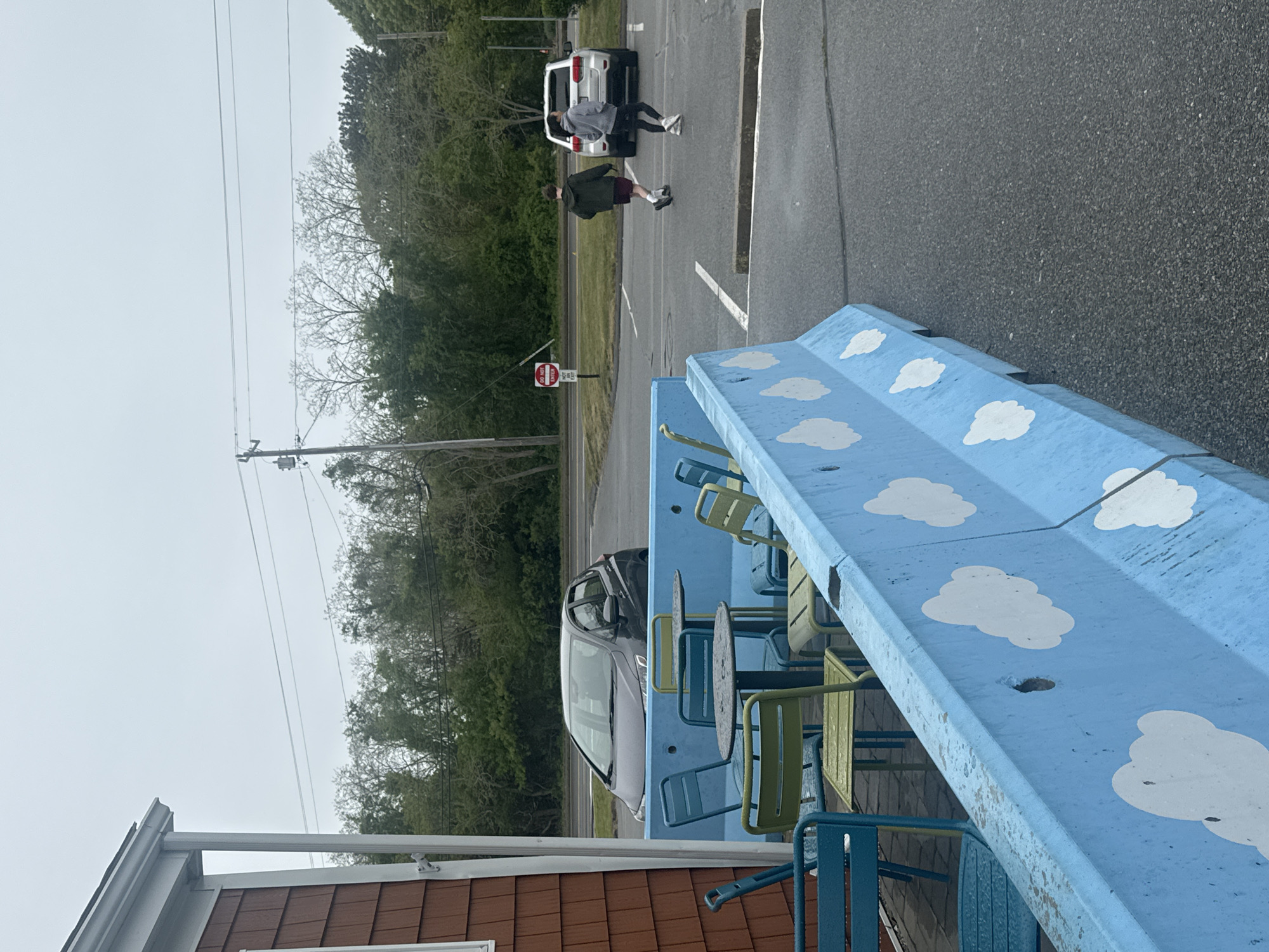The image shows an outdoor scene in what appears to be a parking lot or roadside area in Eastham, United States of America. In the foreground, there is a light-blue concrete barrier decorated with white cloud designs. Behind the barrier are several metal tables and chairs, painted in shades of blue and green. A building with brown siding is visible on the left.
In the background, there's a paved road with cars parked on either side. Two people are walking in the direction of a parked white SUV. A "DO NOT ENTER" sign is visible beside the road. The environment includes trees and utility poles, and the sky is overcast. The asphalt is dark grey.