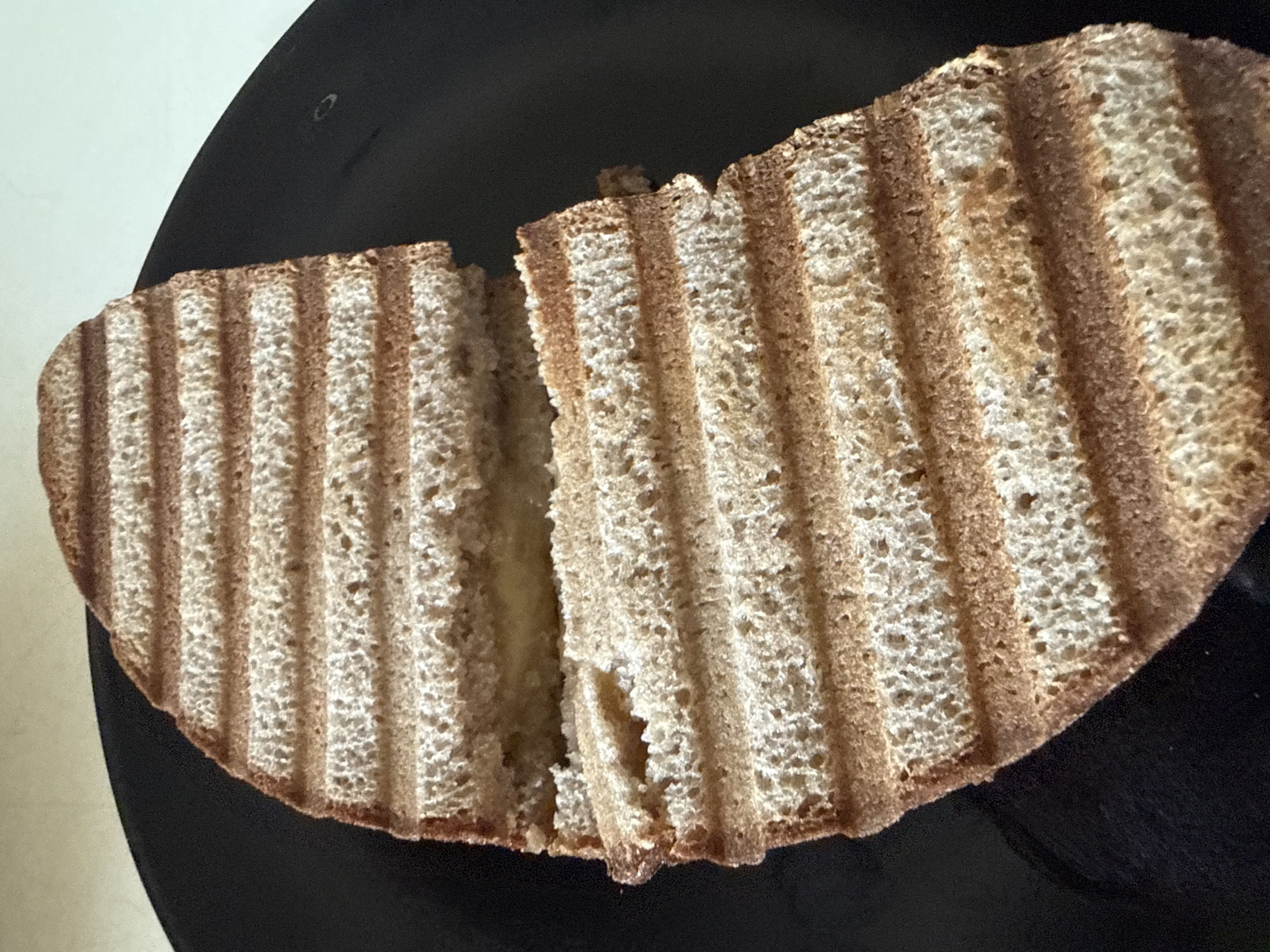 An overhead, close-up shot shows a half-eaten grilled sandwich on a black plate. The sandwich is cut in half vertically and the two halves are slightly apart, revealing a pale yellow filling. The sandwich is made with whole wheat bread that has prominent grill marks. The plate is round and appears to be made of ceramic or plastic. The background is a light beige color. The image is well-lit and the details of the sandwich are clear.