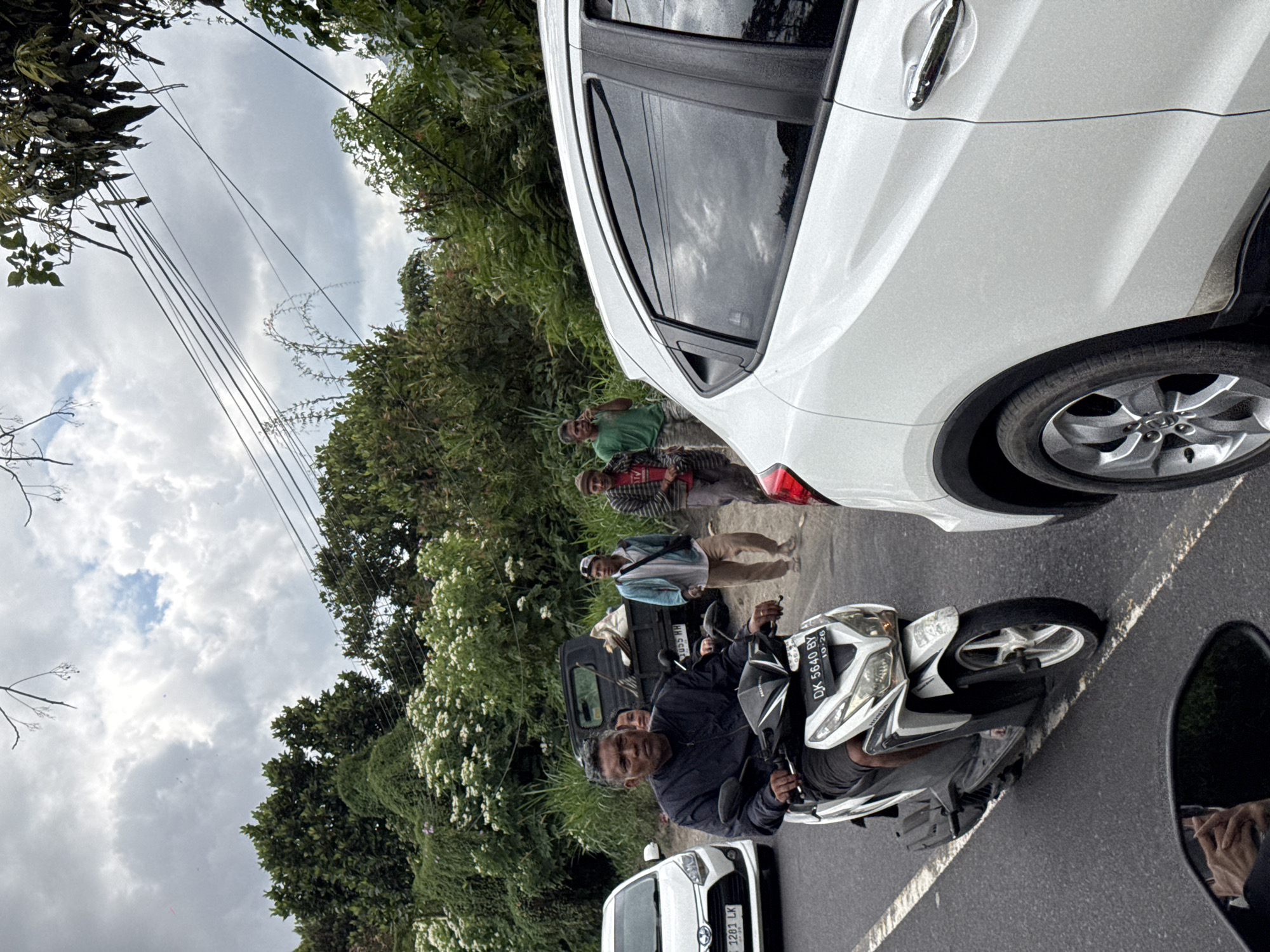 The image depicts a roadside scene in Kintamani, Indonesia, characterized by lush green vegetation and overcast skies. In the foreground, a man is riding a white scooter, wearing a black jacket and dark shorts. The scooter displays the license plate "DK 5640 BY". To the left, a white car with license plate "DK 1281 LK" is visible. Behind the scooter, a dark-colored truck is parked, with its bed appearing to be partially filled with debris or goods. Further back, three men are standing on the side of the road. One man, wearing a light blue jacket and light brown pants, is looking towards the left. Another man, in a green t-shirt, is holding a mobile phone to his ear. The third man, wearing a patterned long-sleeved shirt and a head covering, is standing to the right. The environment suggests a rural or semi-rural setting, with dense foliage bordering the road and visible electrical wires overhead.