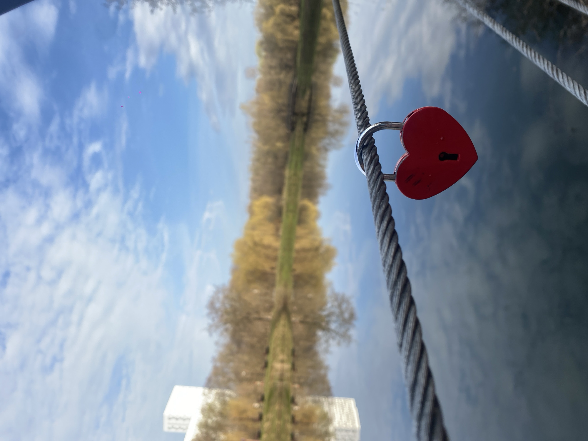 A close-up, eye-level, still shot presents an inverted reflection of an urban environment, specifically Moscow, Russia. A red, heart-shaped padlock is attached to a thick, gray, braided steel cable that runs vertically across the right side of the image. The reflection shows a treelined thoroughfare. The sky is light blue with scattered white clouds. At the bottom of the inverted image, a modern white building can be seen.