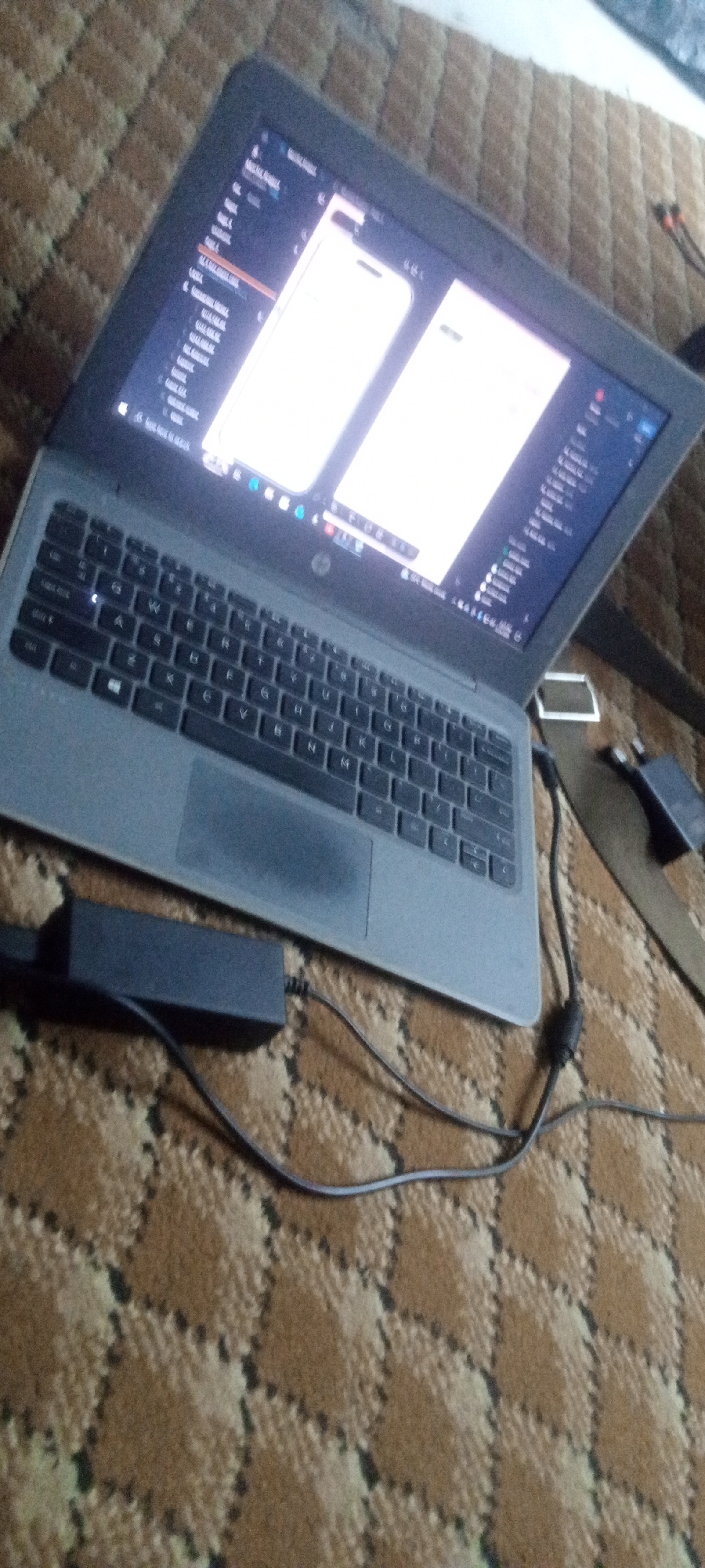 A laptop is sitting on a brown, patterned surface, possibly a carpet or a couch. The laptop is connected to its power adapter. The screen is on and displays two light-colored windows. The location is unknown.