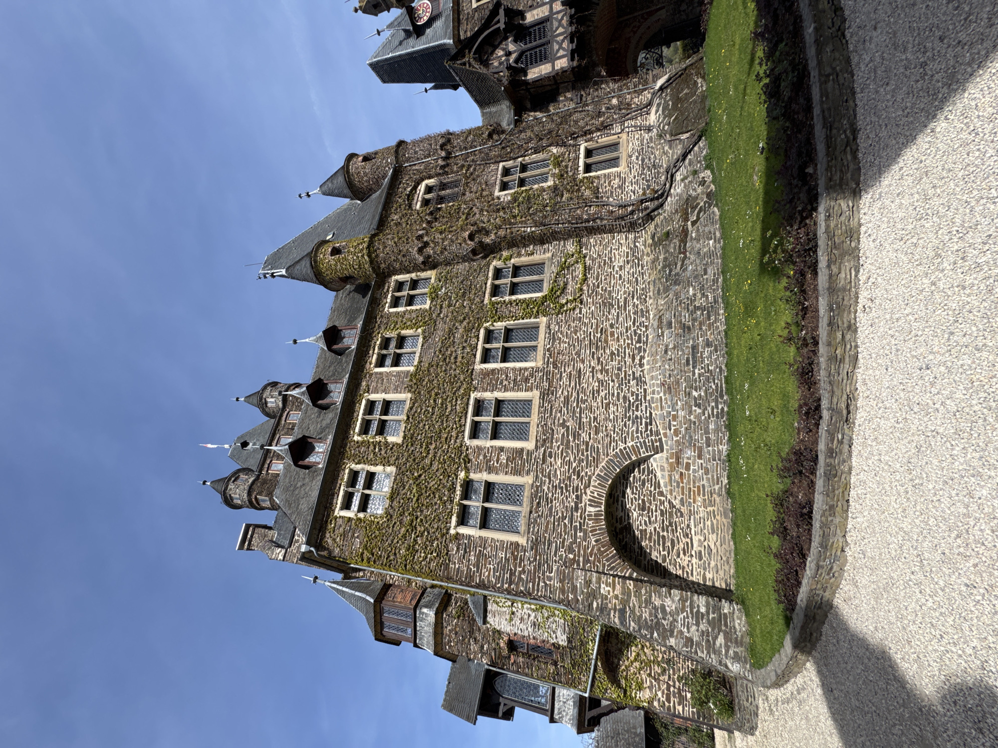 The image shows a building in Cochem, Germany, likely Reichsburg Cochem (Cochem Imperial Castle). The castle is constructed of stone, with multiple towers and turrets visible against a clear blue sky. The walls are partially covered in ivy. Windows are present in rows on the castle walls. At the base of the structure, there is a section of green grass bordered by stonework. Adjacent to the stonework is a gravel path or walkway.