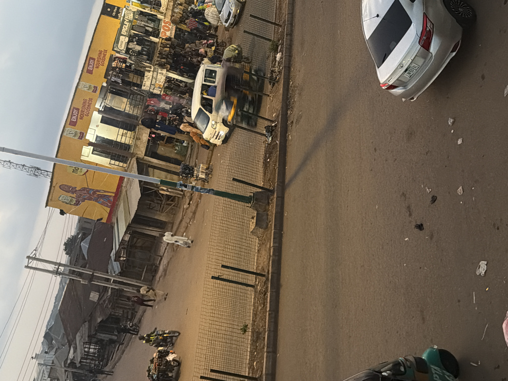 The image depicts a street scene in Bukuru, Nigeria. The street is paved with asphalt and lined with various market stalls and buildings. In the foreground, a silver sedan is parked on the right side of the road. To the left, a three-wheeled vehicle, possibly a tricycle or tuk-tuk, is partially visible. In the middle ground, a white van is in motion, and a person is visible inside it. Several market stalls are set up along the side of the road, displaying various goods. Some people are walking in the street and in front of the stalls. A wire fence runs along the side of the road, separating it from a raised dirt area. Overhead, power lines are strung between poles. The buildings in the background appear to be constructed from corrugated metal and concrete, with some displaying advertisements. The sky is overcast. The overall impression is of a busy, active market area in an urban setting.
