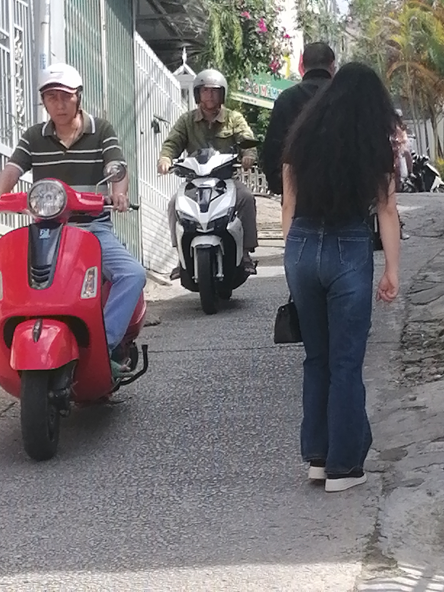 The image depicts a street scene in Da Lat, Vietnam, characterized by its overcast lighting and urban infrastructure. In the foreground, a person wearing a green and white striped polo shirt and blue jeans is riding a red Vespa scooter. This individual is also wearing a white helmet. Behind them, another person, also wearing a helmet and a green jacket, is riding a white motorcycle. To the right, a woman with long, dark hair and blue jeans is walking away from the camera, carrying a black handbag. She is wearing white sneakers. The background features a tall, light-colored metal fence and some foliage, including palm trees and flowering plants. The street surface is asphalt. The overall impression is one of everyday life on a city street.
