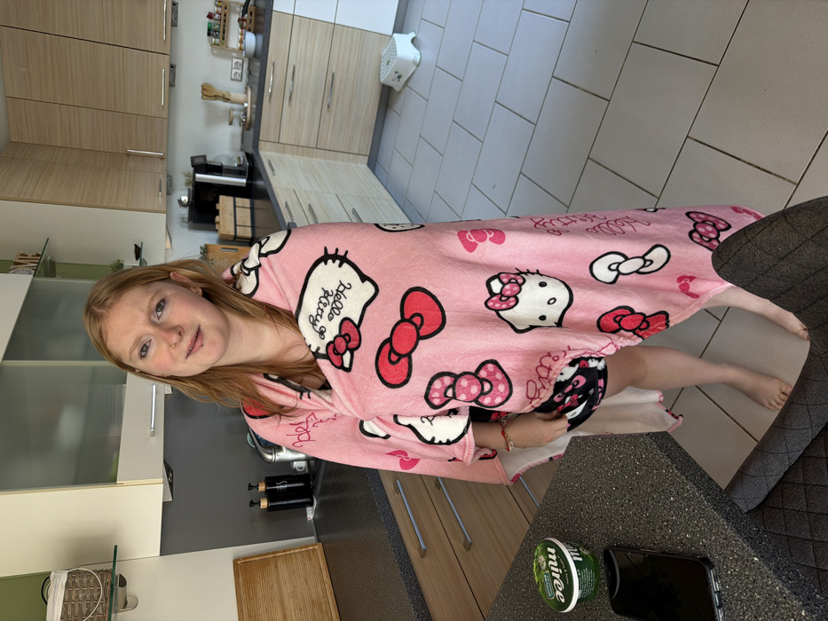 A young adult female with reddish-blonde hair and piercings on her nose and upper lip stands barefoot in a modern kitchen in Trassem, Germany. She is wrapped in a pink Hello Kitty fleece blanket, partially covering black and white patterned shorts. She faces forward, looking directly at the camera with a slight, neutral expression. The kitchen features dark gray speckled countertops, light wood-toned base cabinets, and white/frosted glass wall cabinets. The floor is covered with large light gray square tiles. On the left countertop, a wooden cutting board, two dark pump bottles, a green plastic container labeled 'Nuinée', and a black smartphone are visible. A dark gray upholstered chair is partially visible in the lower foreground. The right section of the kitchen displays a dark coffee machine, a wooden storage box, and a tiered spice rack on the counter. A small white plastic step stool with a green floral design rests on the tiled floor near the far wall.