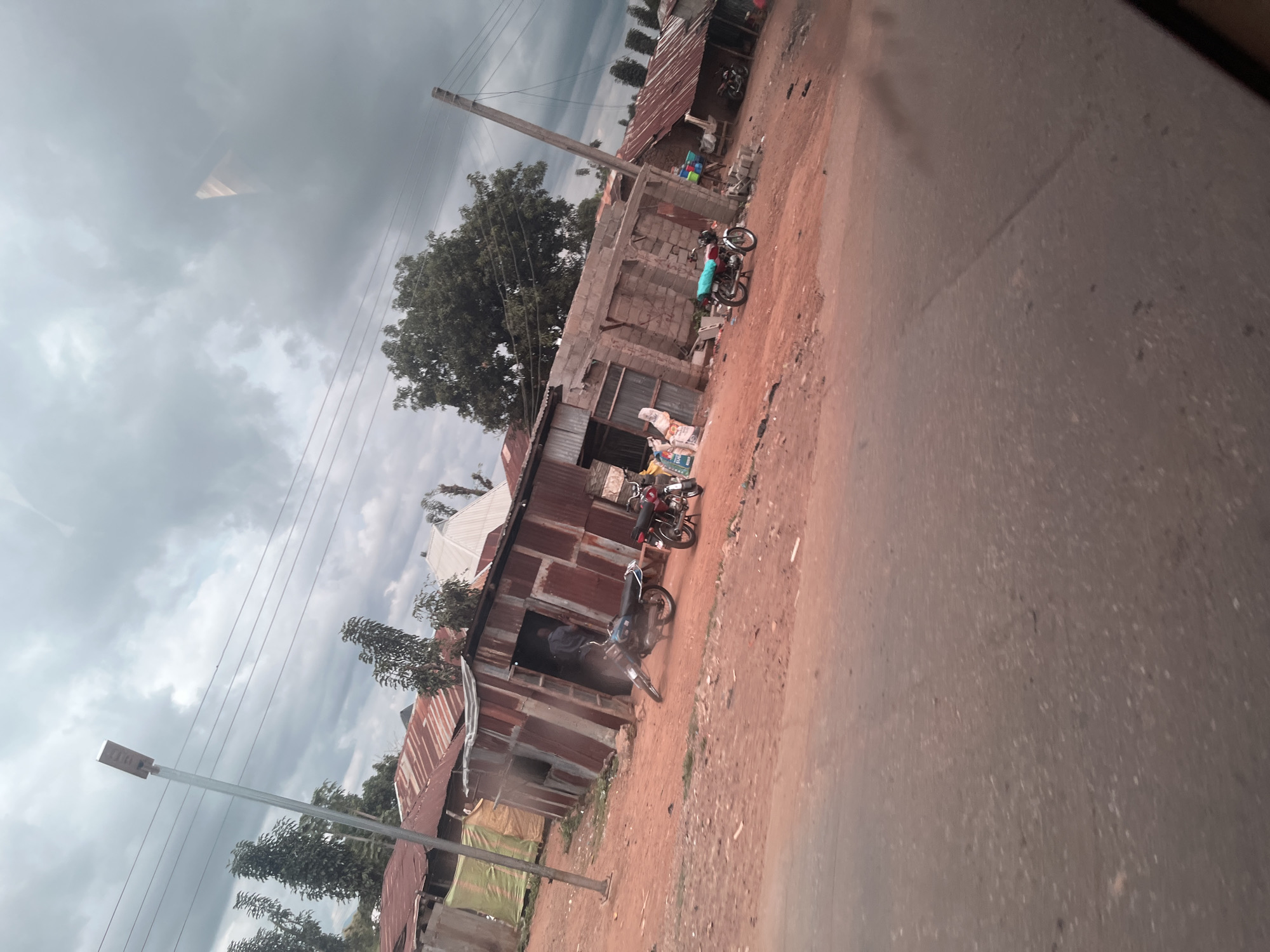The image depicts a street scene in Karshi, Nigeria, under a cloudy sky. The foreground shows a section of a dirt road, blurred due to motion, with red earth and scattered debris. Beyond the road, a collection of structures, likely shops or homes, are visible. These buildings are constructed with corrugated metal and, in some cases, cinder blocks, featuring rusty roofs and open entrances.
Several motorcycles are parked in front of the buildings. In one doorway, a person in a blue shirt is partially visible, appearing to be inside a shop. Bags of goods are stacked near some of the structures. A modern solar-powered street lamp stands prominently on the left side of the image, with electrical wires stretching across the sky. Trees with dark green foliage are scattered throughout the scene. The overall impression is one of a functional, possibly informal, commercial or residential area.