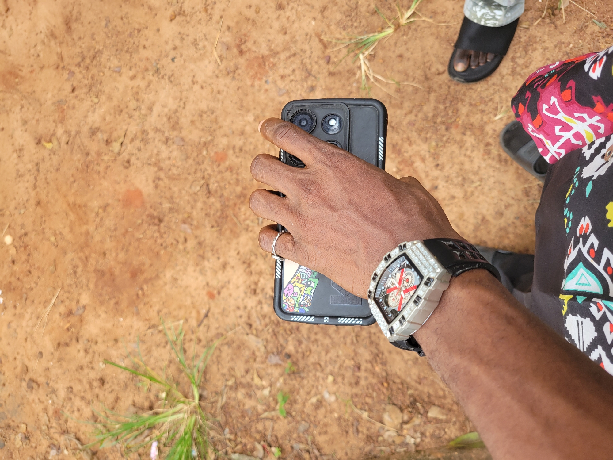 A person's hand is holding a black smartphone with a colorful patterned case and a bezel-set diamond watch on their wrist. The phone is held at eye level, pointing downwards towards the ground. The background is a reddish-brown dirt path with sparse green grass and dry twigs scattered across it. Two black sandals are visible on the ground, one to the right and another partially visible to the left, suggesting the person is sitting or resting on the ground. The scene is likely outdoors in a rural or semi-rural area, possibly the city of Ukukwa, Nigeria, given the reddish soil and vegetation.