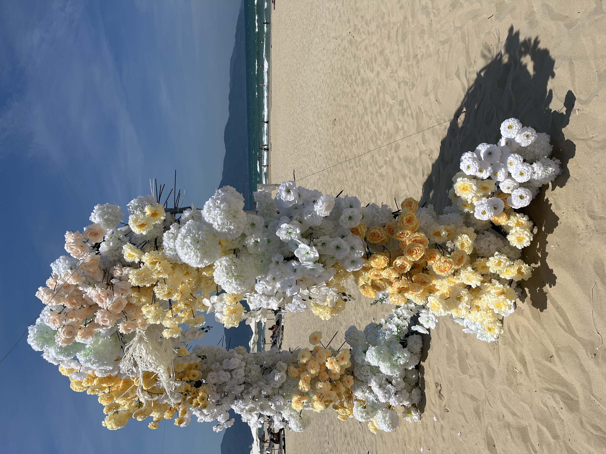 The image captures a picturesque beach scene, showcasing an elaborate floral arrangement likely prepared for a special event. In the foreground, a large, partially visible structure, appearing to be a heart-shaped or circular arch, is richly adorned with artificial flowers. The blooms are predominantly in shades of white, cream, pale yellow, and light peach, arranged in dense clusters resembling hydrangeas and individual rose-like blossoms. White macrame-style tassels hang delicately from sections of the floral decor, and black support sticks are discernible within the arrangement.
The floral structure is positioned on a wide expanse of light-colored sandy beach, which displays subtle textures and ripples. Strong, distinct shadows are cast on the sand by the floral arch and possibly the photographer, indicating bright, direct sunlight, suggesting it is mid-morning or early afternoon. Beyond the immediate foreground, the tranquil ocean stretches out, with gentle waves breaking onto the shore. Small, indistinct figures of people are visible in the water and walking along the coastline, enjoying the natural setting. In the distant background, majestic mountains rise, framing the scene against a clear, vibrant blue sky with a few faint, wispy clouds.
The overall scene strongly suggests a beautiful beach wedding ceremony, engagement proposal, or another celebratory event. The location, with its sandy beaches, ocean, and mountains, aligns perfectly with coastal regions like Đà Nẵng, Vietnam. The atmosphere conveyed is one of romance, joy, and natural splendor. There is no visible text in the image.