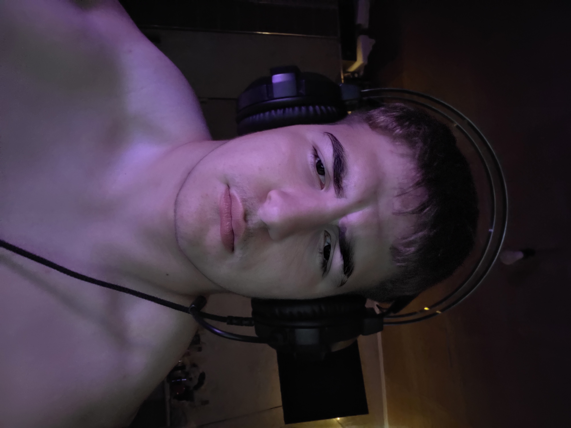 The image shows a close-up, chest-up view of a young male wearing headphones. He is shirtless and illuminated by purple lighting. The background is dark and indistinct, with hints of furniture and a screen on the right. No specific location in Aleksandrovo, Bulgaria is discernible from the image. The man's expression is neutral to slightly concerned. The focus is on his face and upper body.