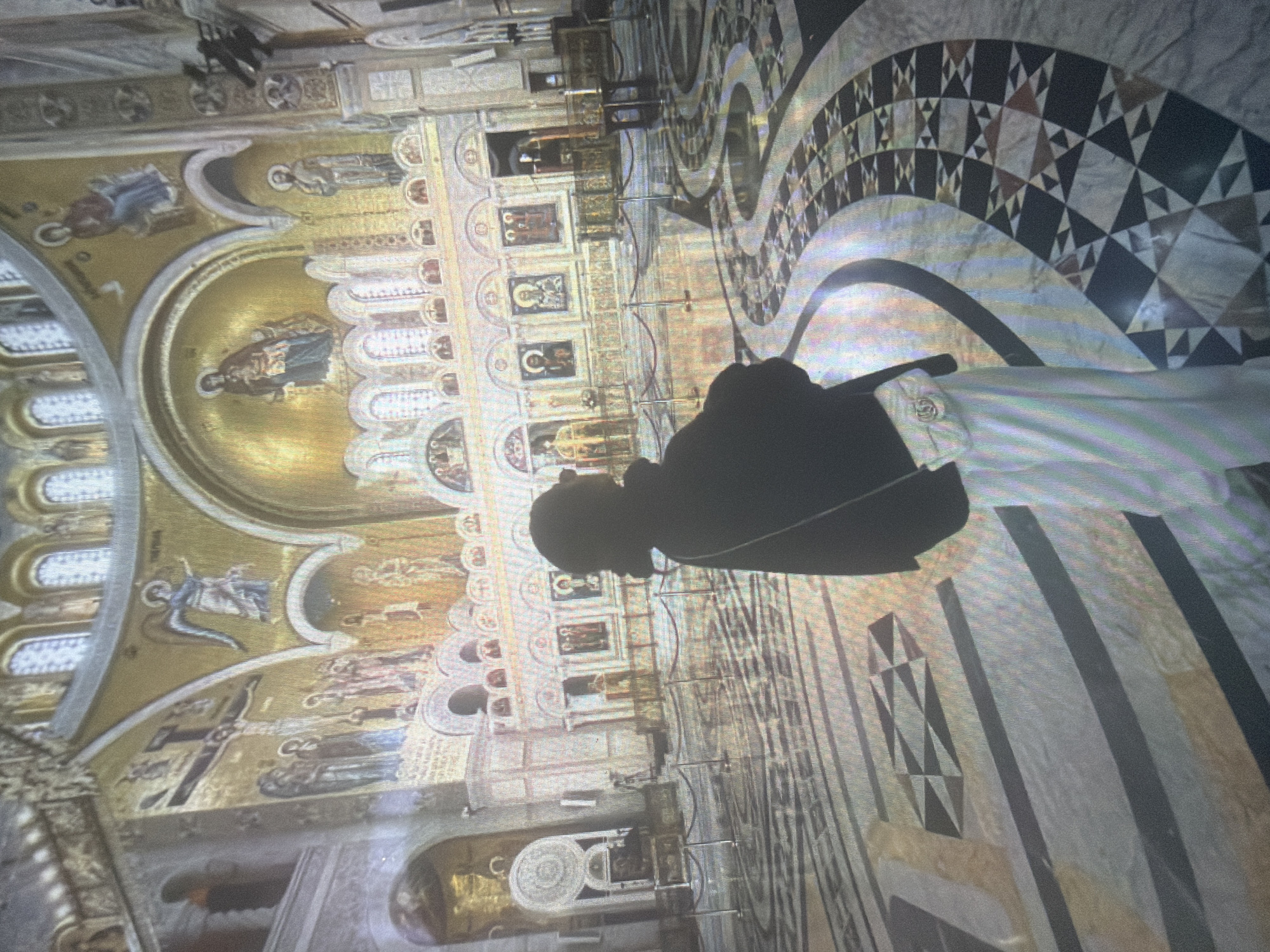 The image displays the lavish interior of the Temple of Saint Sava in Belgrade, Serbia, not Shumen, Bulgaria. A single individual stands with their back primarily to the camera, positioned slightly right of center, observing the expansive hall. The person is dressed in a dark top, light-colored wide-leg pants, and carries a small white shoulder bag featuring a visible Gucci logo. Their head is rendered in deep silhouette. The upper walls and domed ceilings are extensively covered with detailed golden mosaics depicting various religious figures and biblical scenes, including Christ Pantocrator in the central dome, an angel with outstretched wings on the upper left, the Virgin Mary, saints, and a crucifixion scene. The architecture features grand arches, windows, and ornate structures. The floor is composed of highly polished marble with intricate geometric patterns in shades of black, white, gray, and earthy tones, creating swirling and angular designs. Red velvet ropes on metal stanchions are set up across the floor, demarcating pathways for visitors.