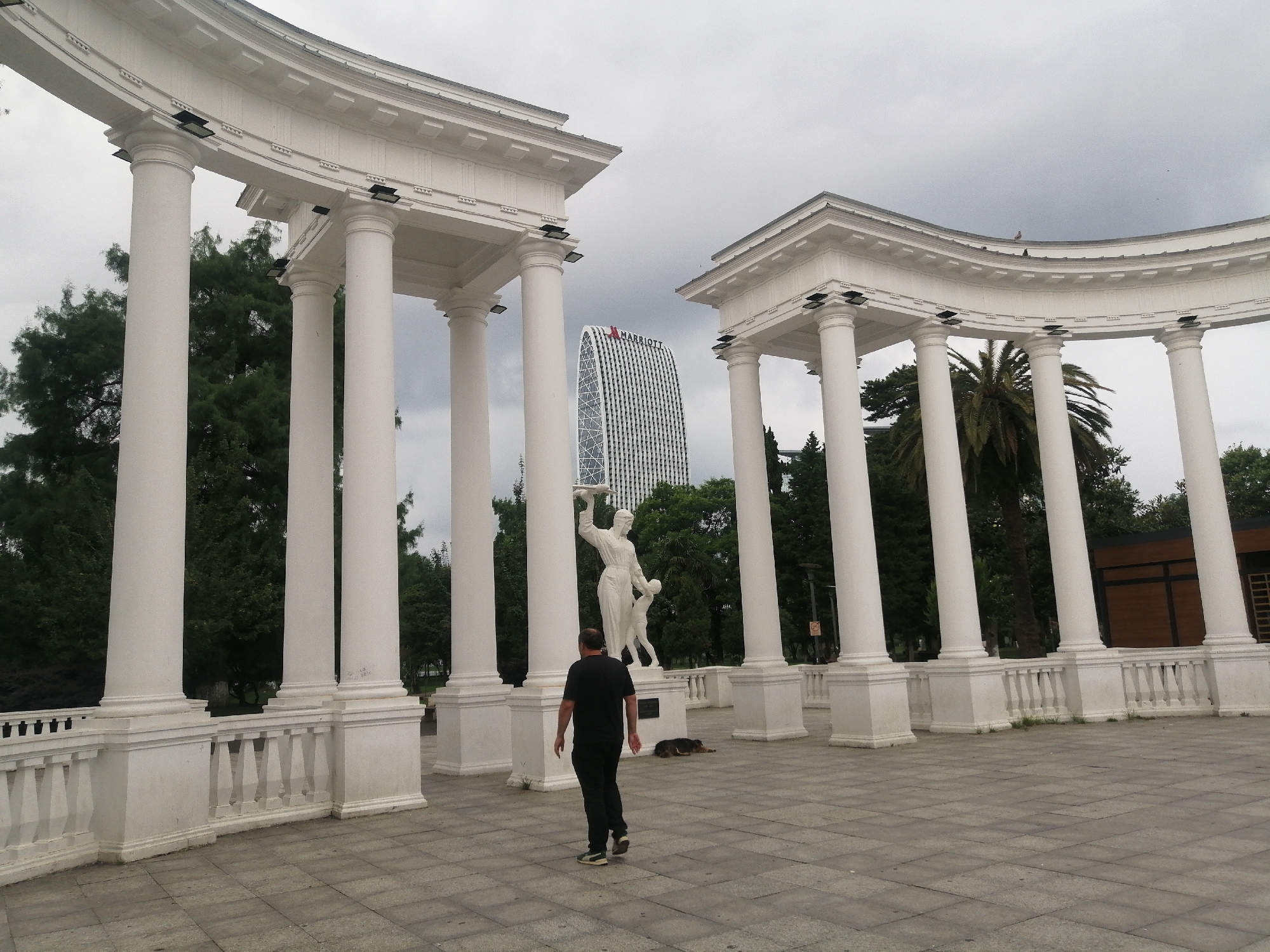 The image shows a man walking away from the camera in a large, open-air plaza in Batumi, Georgia. He is wearing a black t-shirt and dark pants. The plaza is paved with gray tiles and features a white, curved colonnade with numerous columns. In the background, a tall, modern skyscraper with the Marriott logo is visible, surrounded by trees. A white statue of a woman holding a platter and a child stands in the middle ground. A dog lies on the ground near the statue. The sky is overcast, suggesting cloudy weather. This location is likely the Alphabet Tower area or a similar public space with neoclassical architecture in Batumi.