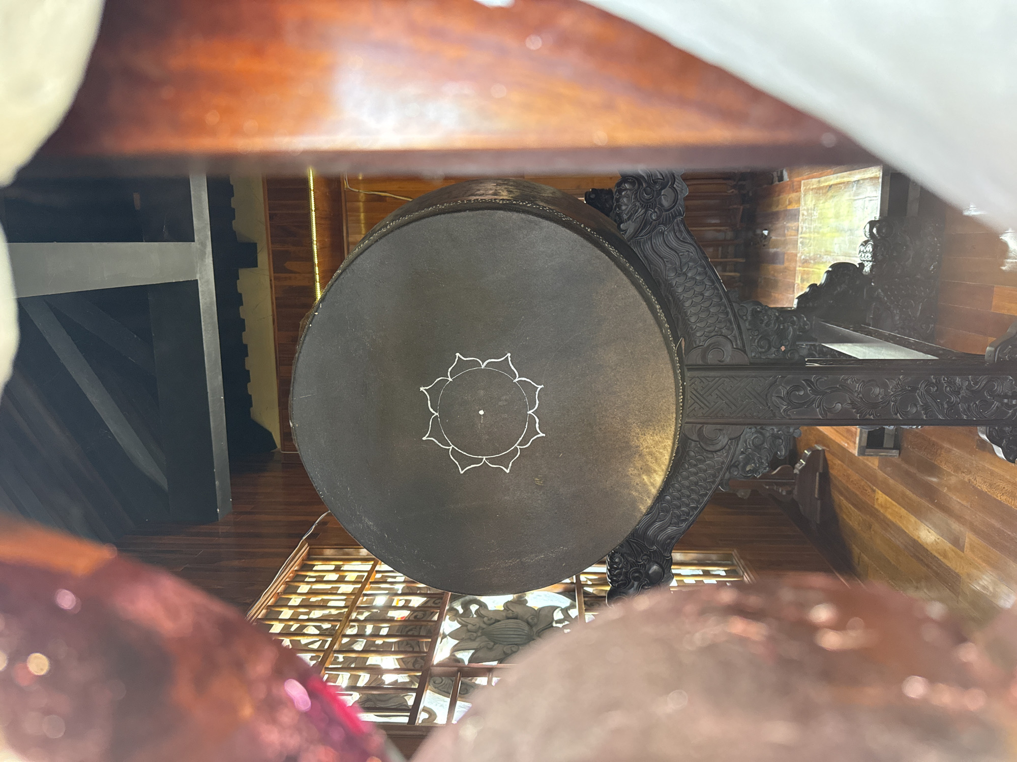 A low-angle shot of a hanging gong, suspended by a carved wooden frame. The gong is round, dark, and features a lotus-shaped design at the center. The wooden frame has intricate carvings, including dragon-like figures. The background includes wooden walls and a gridded ceiling, possibly part of a traditional building. The location is Tay Nha Trang Ward, Vietnam.