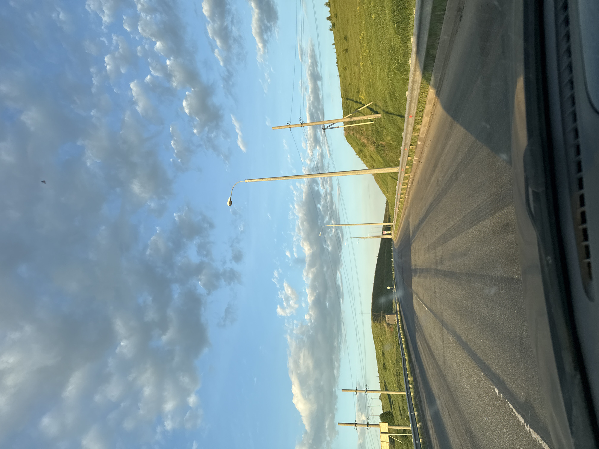 The image presents a view from inside a vehicle traveling along an open asphalt road in what appears to be a rural or semi-rural area, likely near Тарасово, Russia. The road, dark grey with visible tire marks and faint white lines, stretches into the distance. A silver metal guardrail with small red/orange reflectors lines the right side of the road, separating it from a gently sloping green hillside covered in lush grass. Similar green embankments and fields are visible on the left side.
Utility poles, both wooden and concrete, with power lines strung between them, line both sides of the road. Modern streetlights with curved arms are also prominent on the left. The sky above is a clear, bright blue, dotted with scattered white and light grey cumulus clouds. A small, dark, bird-like object is faintly visible in the upper left sky.
The lighting suggests late afternoon or early evening, with the sun casting long shadows, particularly from the guardrail onto the road, indicating a low sun angle. The weather appears fair and sunny with light cloud cover. The overall scene depicts a peaceful journey through an open, green landscape. No people or visible text are present in the image.